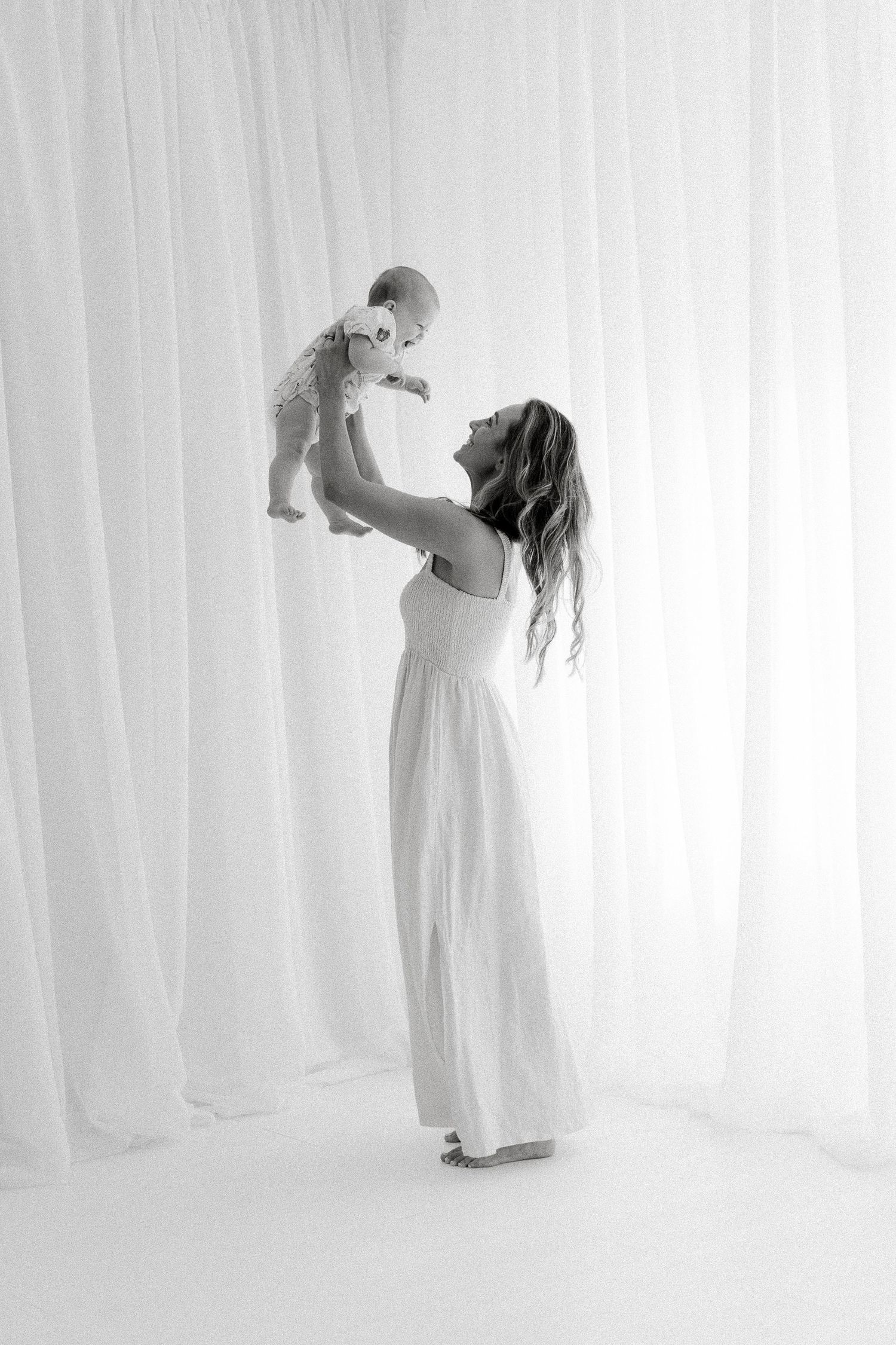 Black and white photo of a mother in white dress lifting her baby overhead, smiling joyfully in a bright studio.