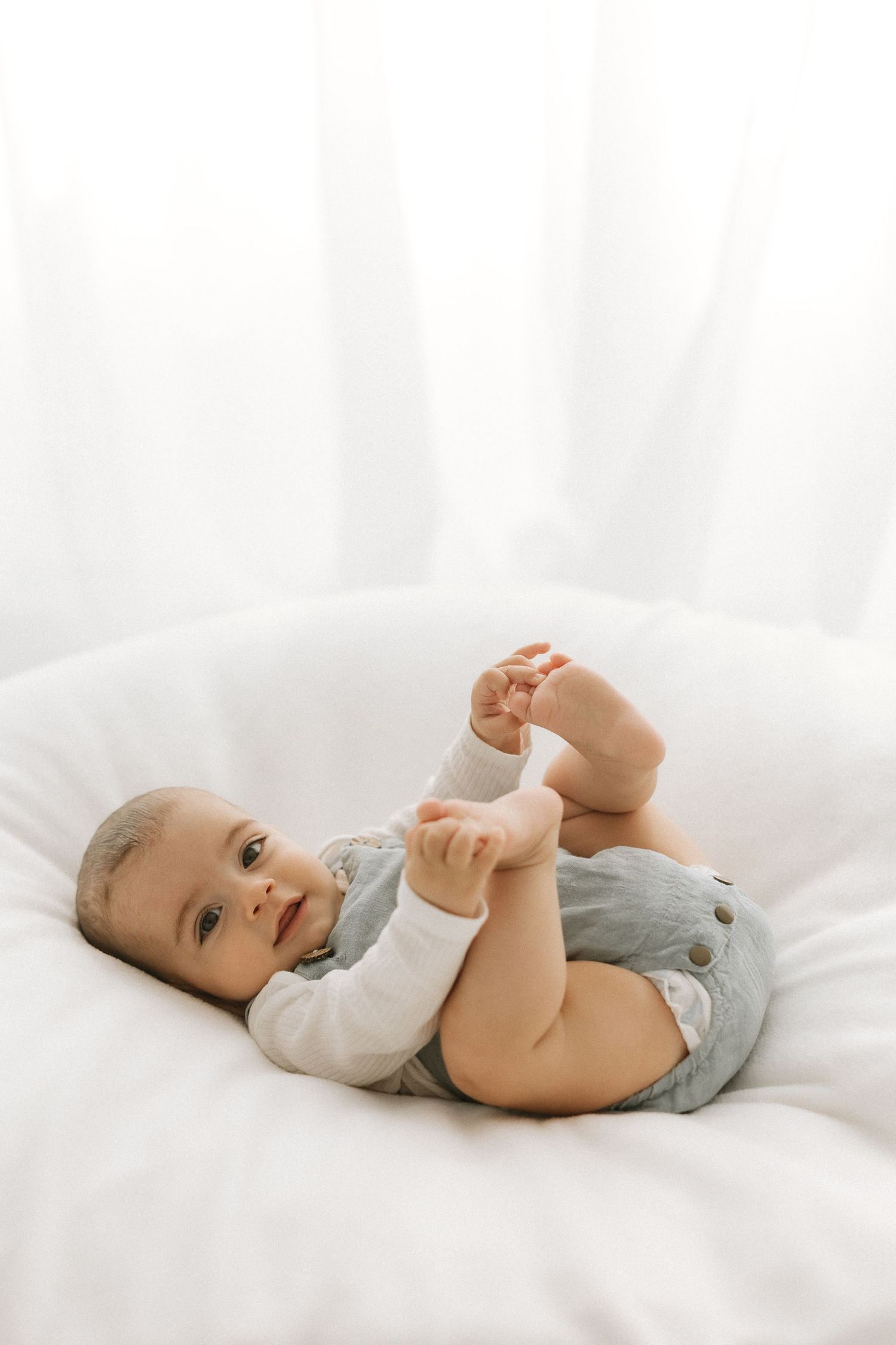 Adorable baby lying on white bedding, holding feet up, wearing gray overalls and white long sleeves.