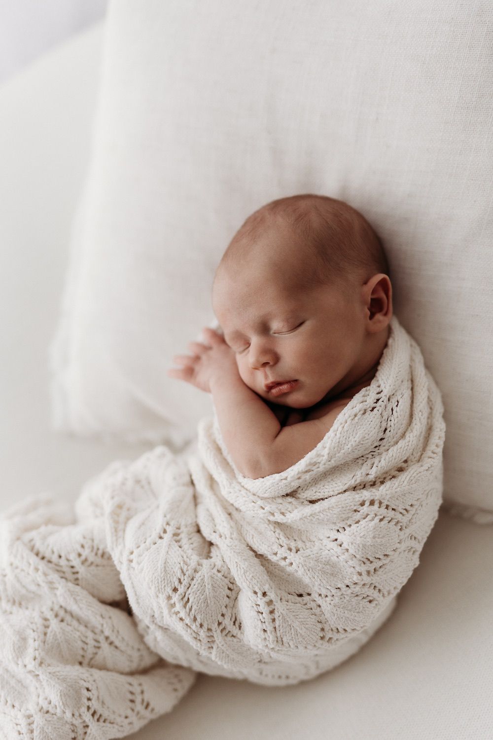 Peaceful newborn baby swaddled in cream knit blanket sleeping against white pillow.