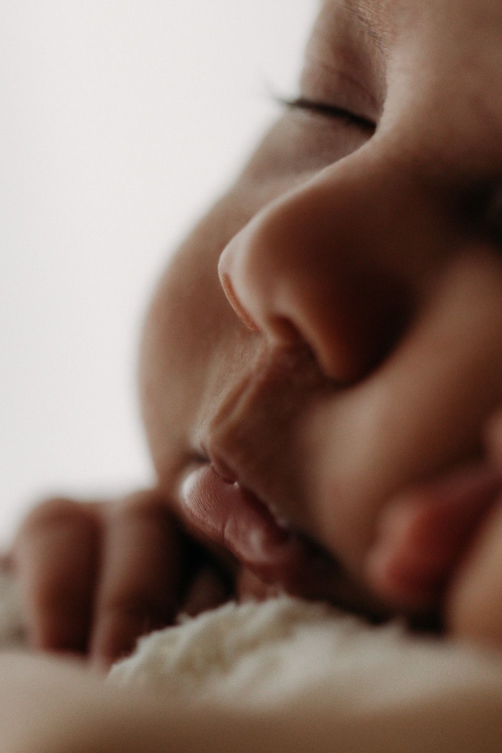 Close-up portrait of a sleeping newborn baby with tiny fingers near soft lips on white blanket.