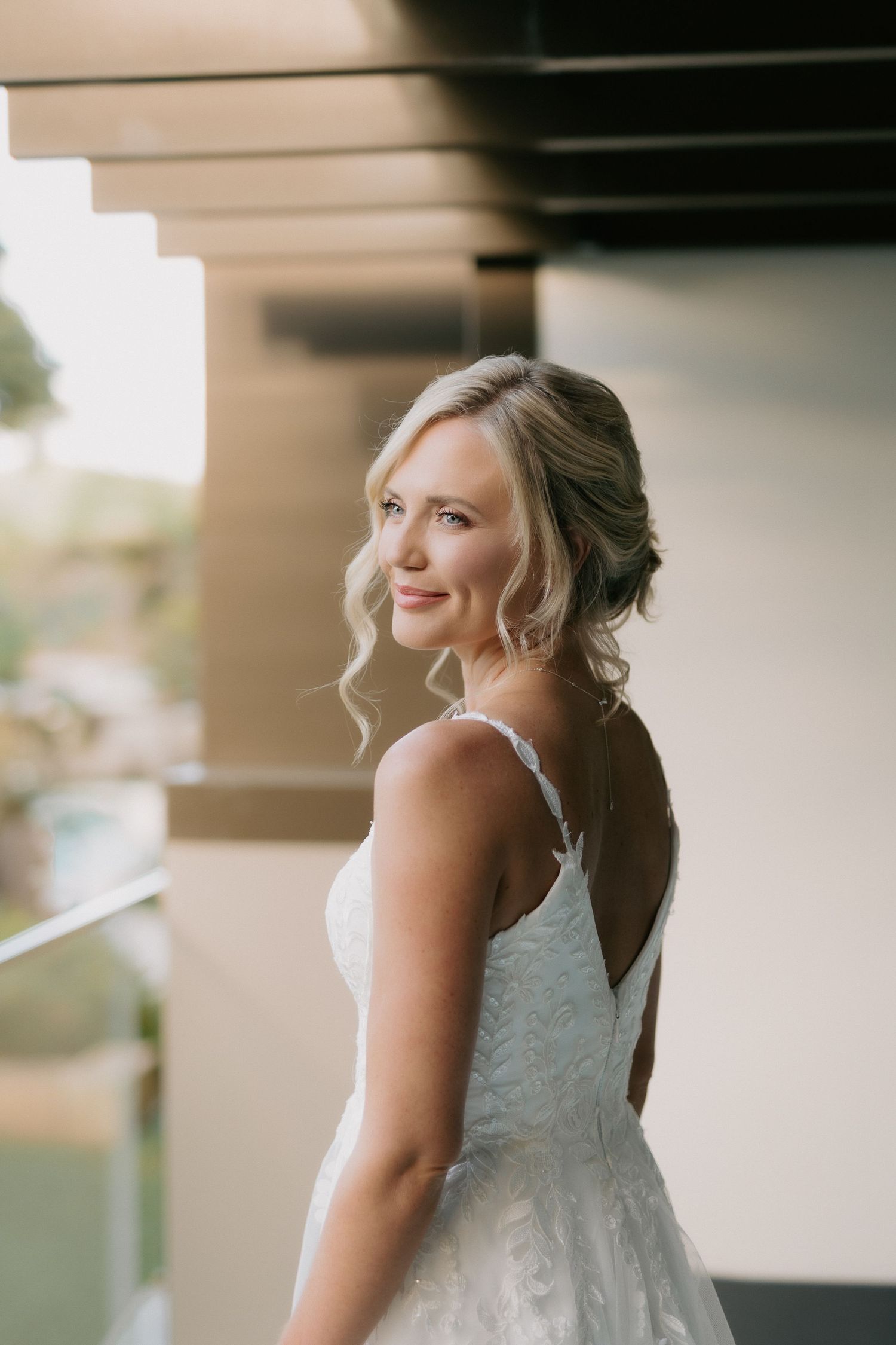 Elegant bride in a backless lace wedding gown smiling over her shoulder on a sunlit balcony at Noosa wedding resort ready for her vows.