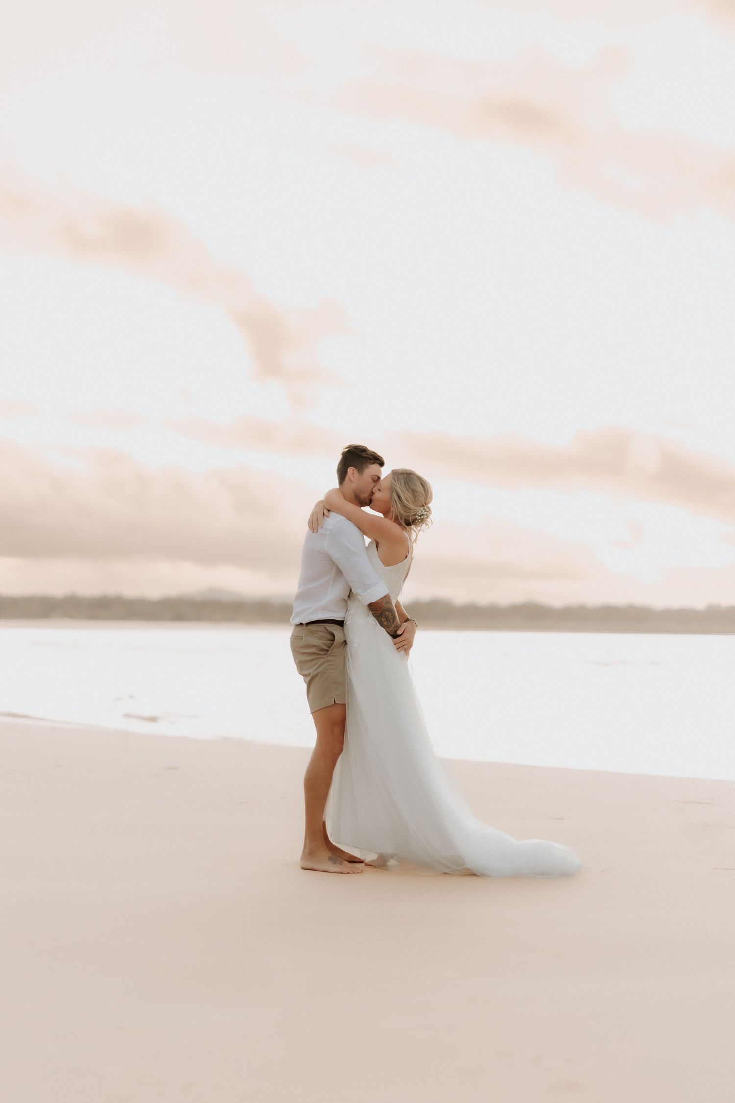 Bride and groom embracing barefoot on a sandy beach at sunset during their wedding day on Noosa beach.