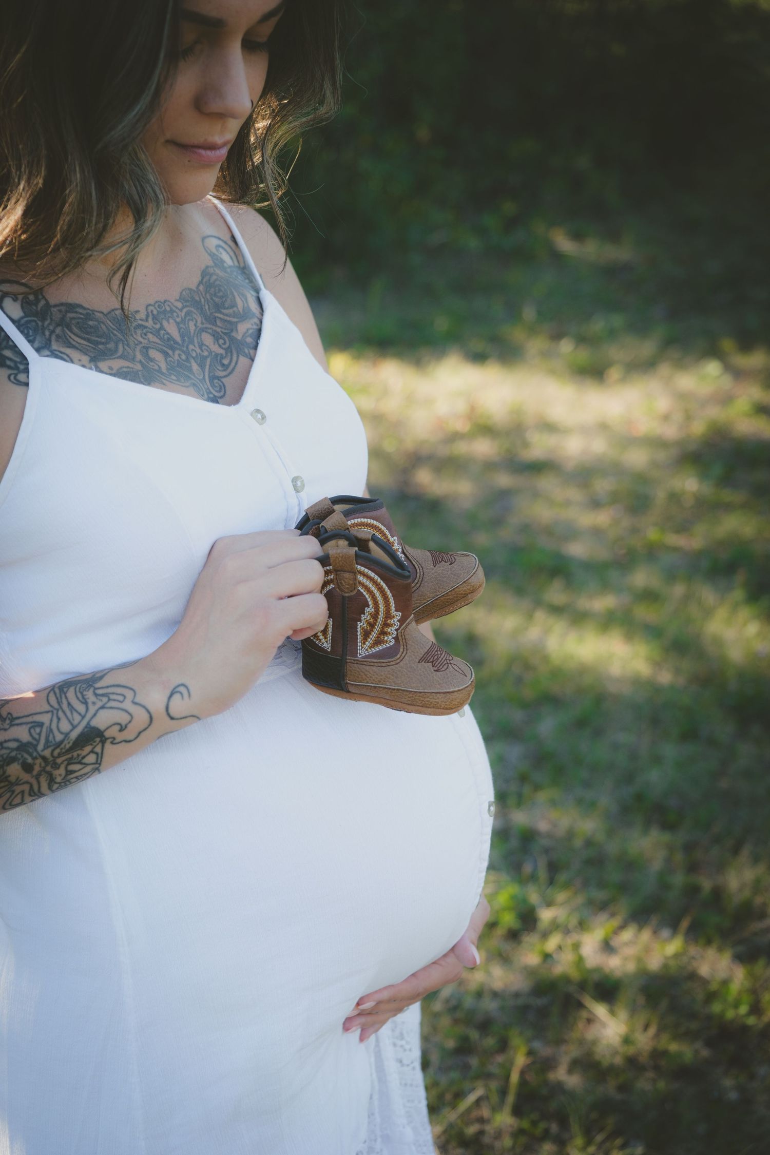 Tattooed pregnant woman in white dress holding tiny baby shoes against her baby bump outdoors.
