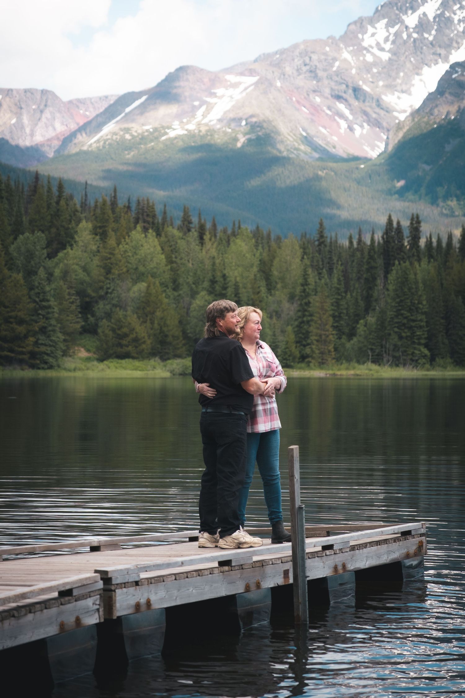 Romantic couple embracing on wooden dock with stunning mountain lake and snowy peaks behind them.