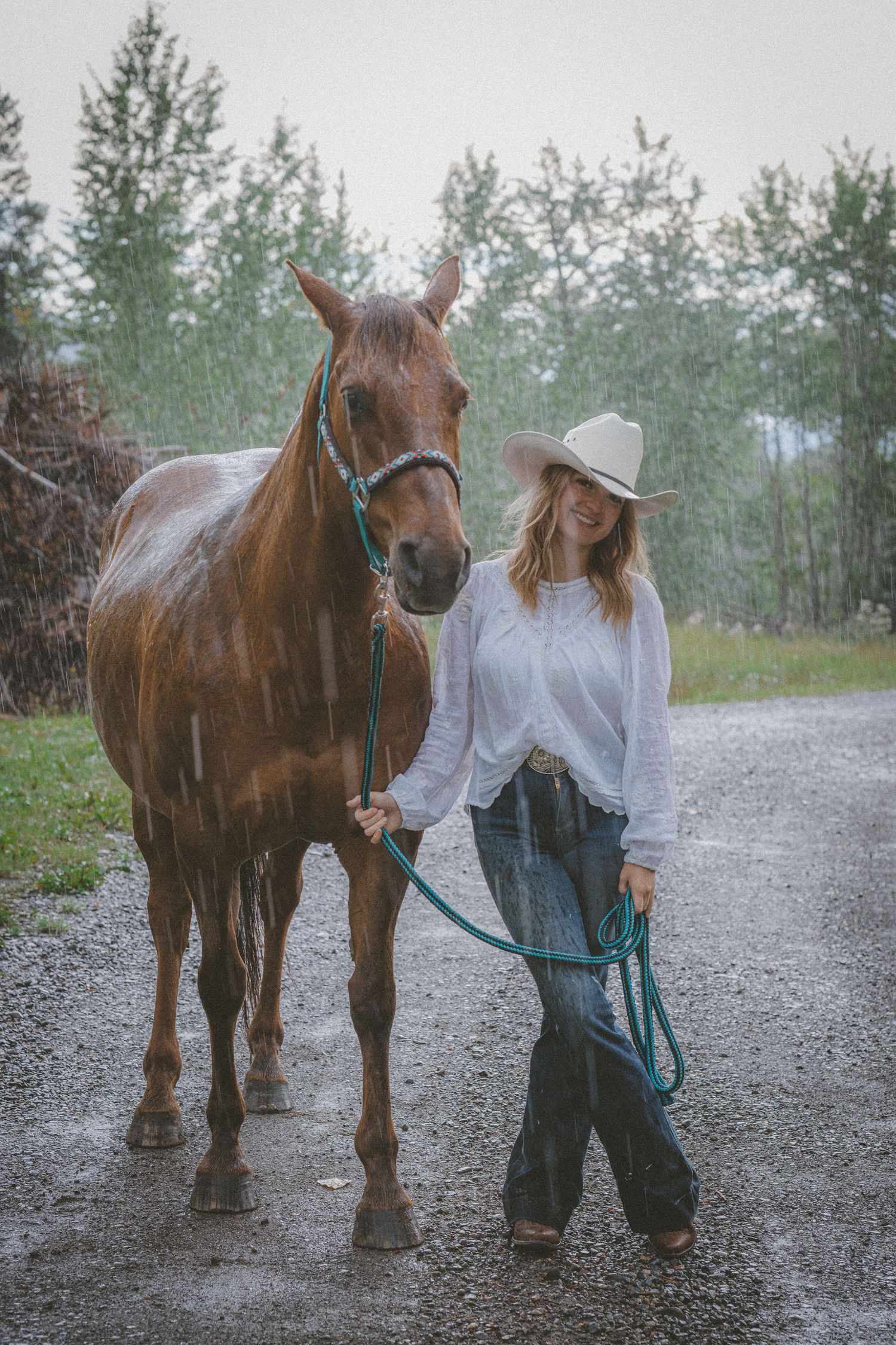 A woman in a cowboy hat and jeans leads a chestnut horse down a gravel path in the rain.