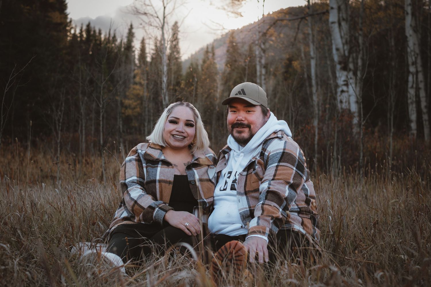 A smiling couple in matching plaid shirts sit together in a golden autumn meadow with mountains behind.