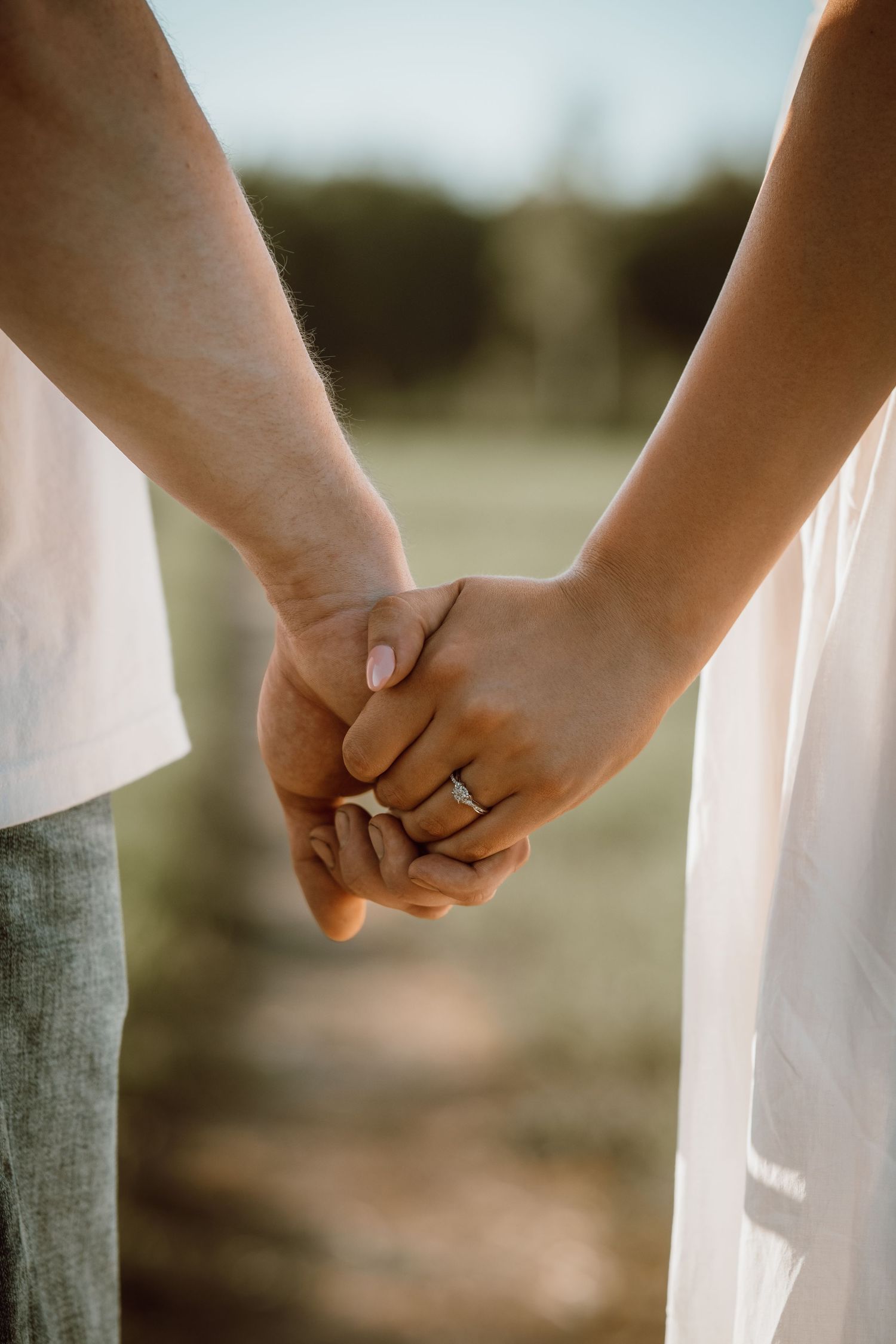 Close-up of a couple holding hands in a field, woman wearing an engagement ring, both in white outfits.