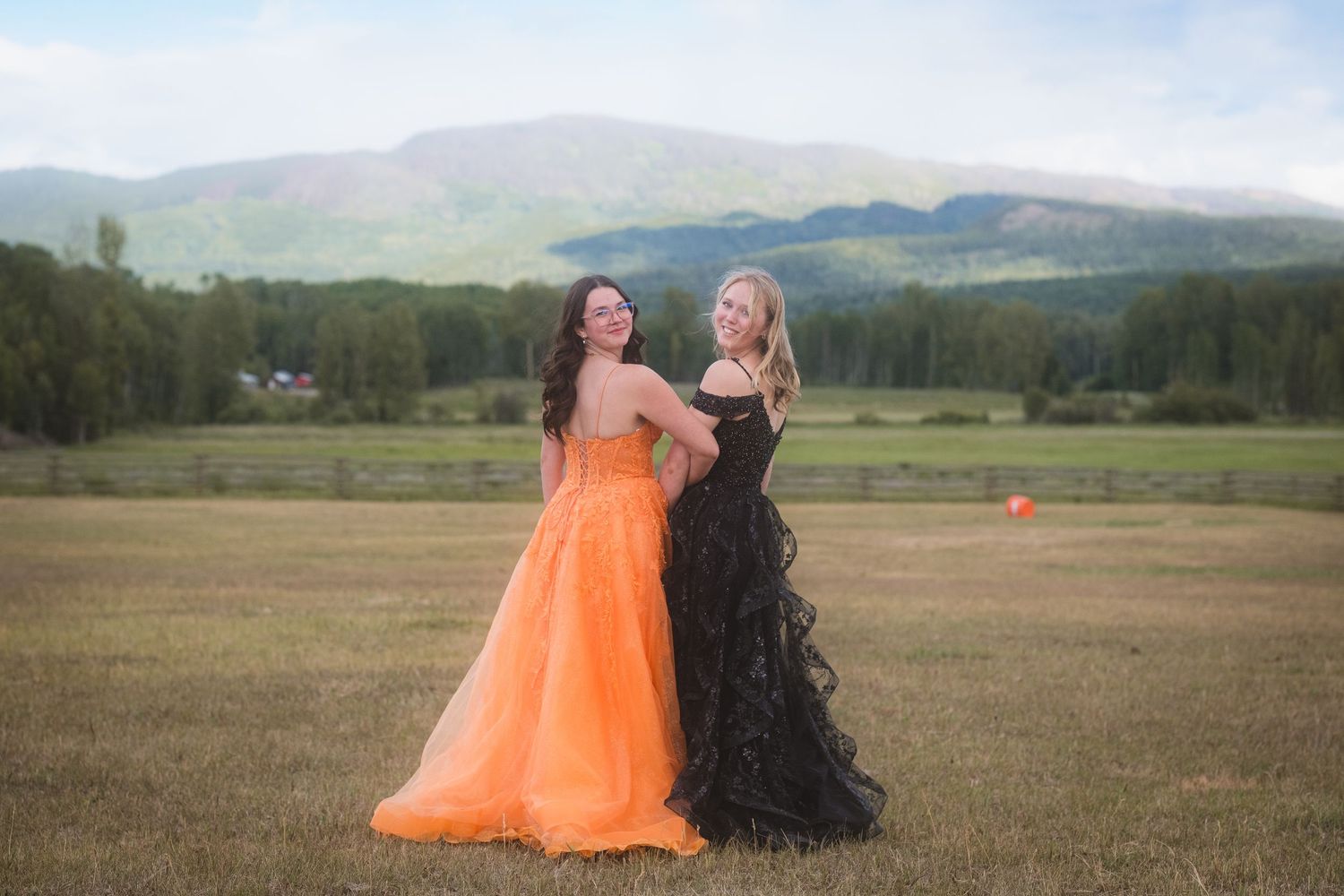 Two young women in orange and black formal gowns pose in field with mountain view.