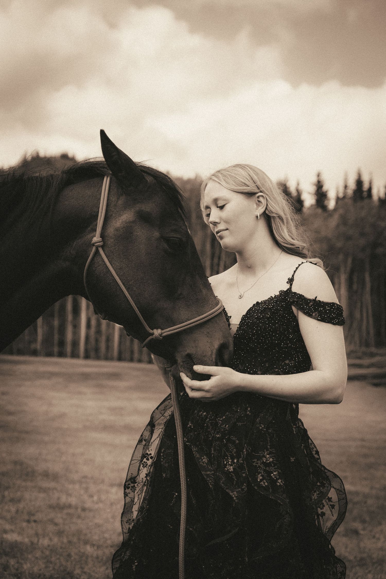 A sepia-toned portrait of a blonde woman in a black lace dress tenderly touching a dark horse outdoors.