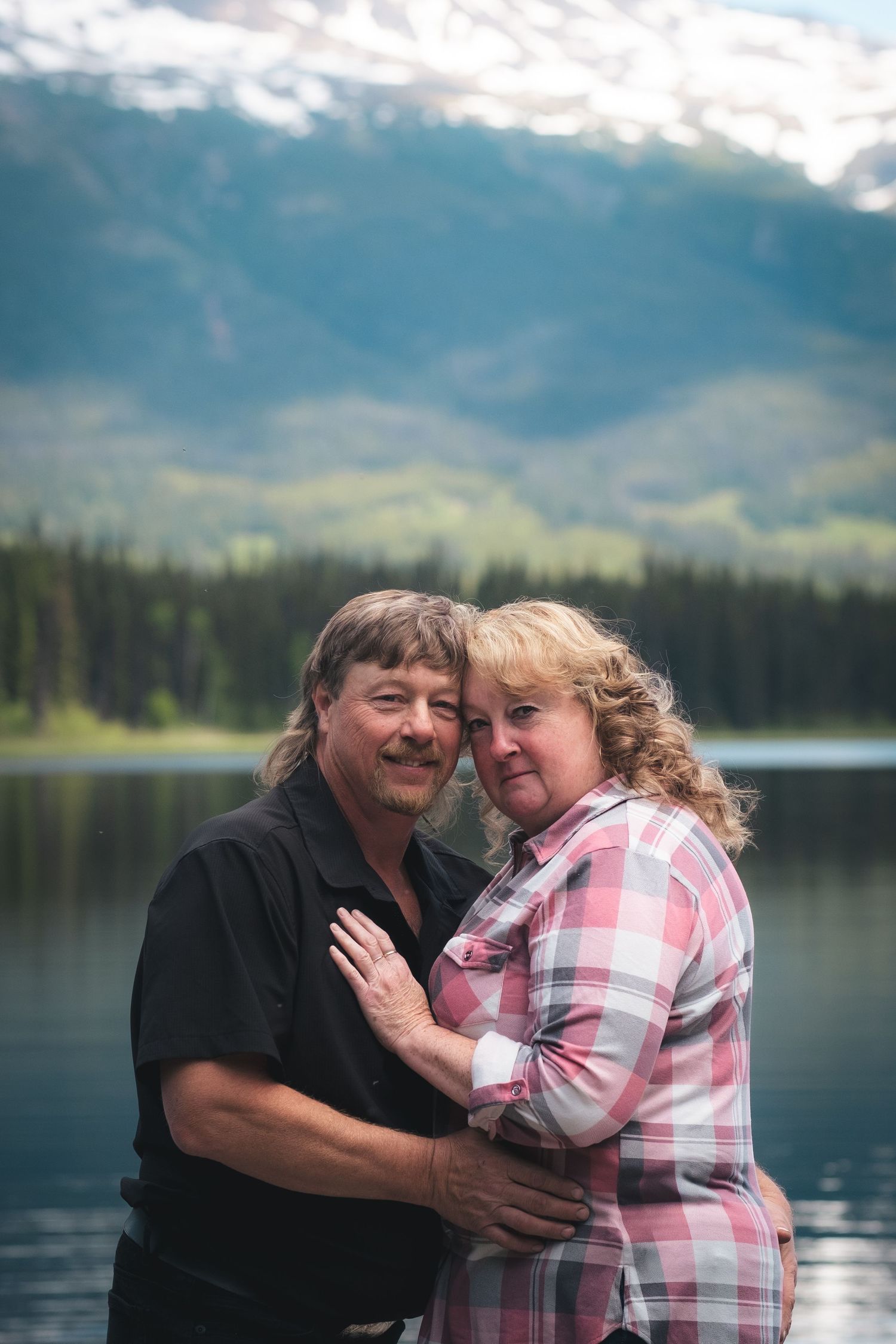 Smiling middle-aged couple embracing with scenic mountain lake and snow-capped peaks behind them.