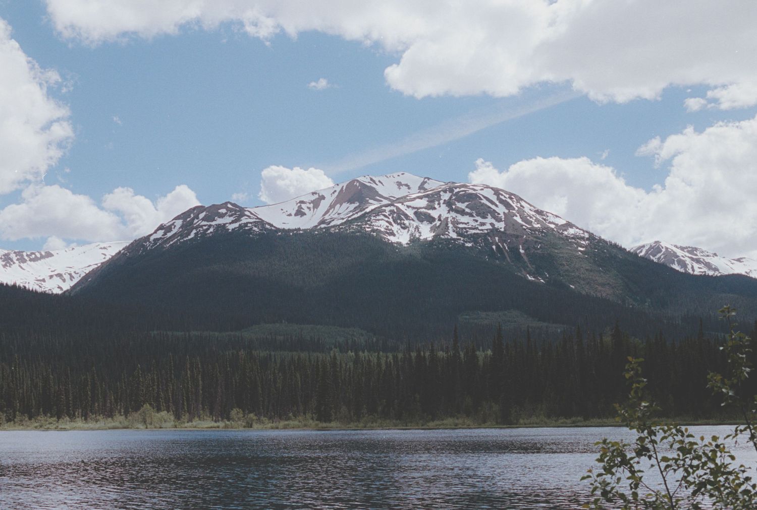 Snow-capped mountain peaks rise above a dense pine forest reflected in a calm alpine lake.