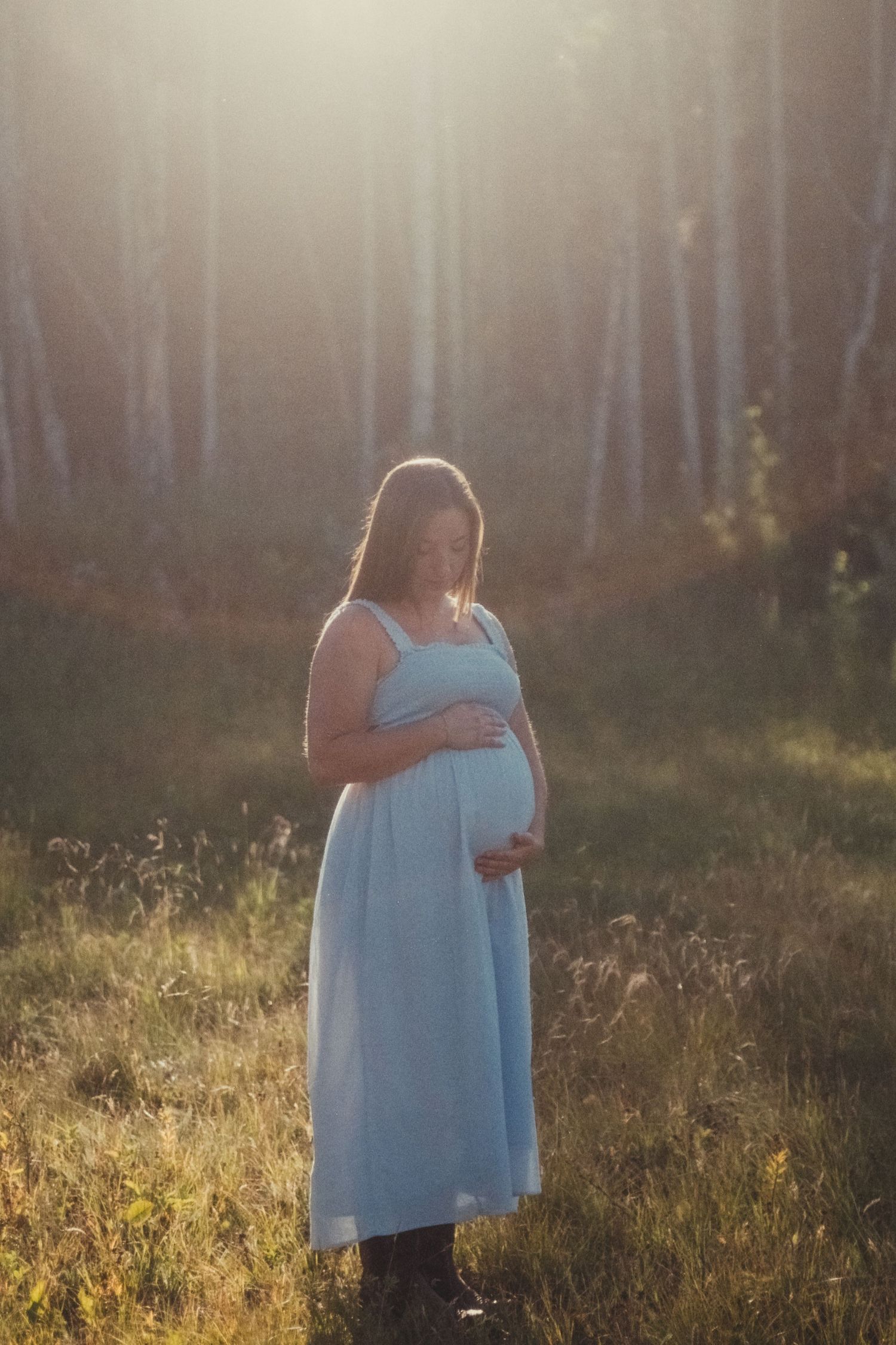 Glowing pregnant woman in light blue dress holding belly in sun-drenched forest setting.