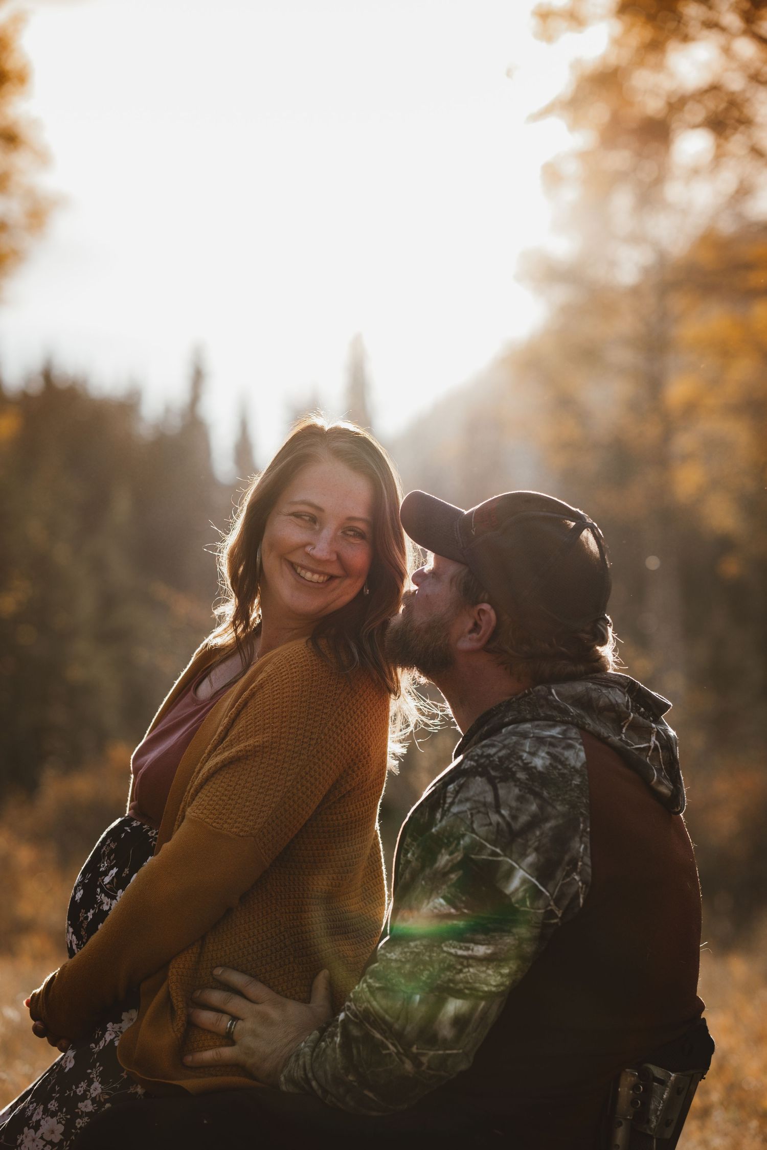 Smiling woman in mustard cardigan being kissed by bearded man in camo in autumn light.