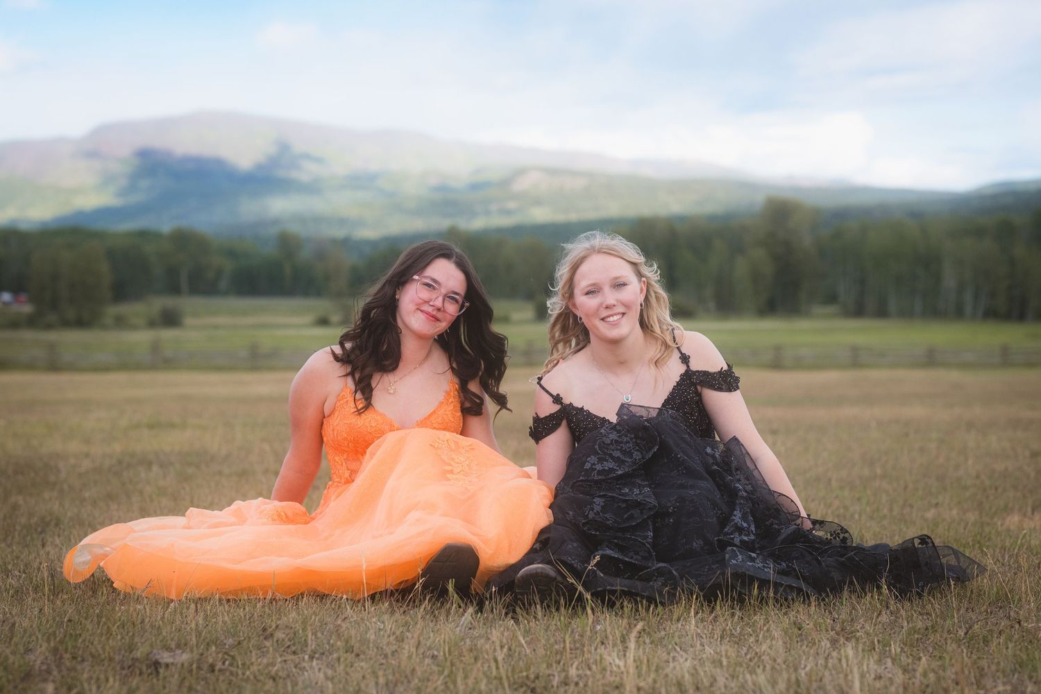 Two young women in formal gowns sitting in a field with mountain views.