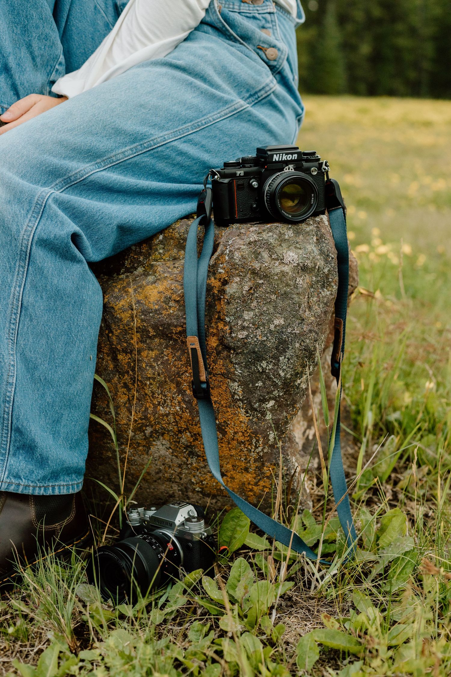 Nikon camera resting on a mossy rock beside a person in denim overalls sitting in a grassy field.
