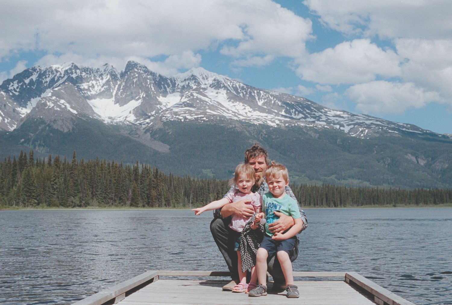 A man and two young children pose on a dock with stunning snow-capped mountains and a lake behind them.
