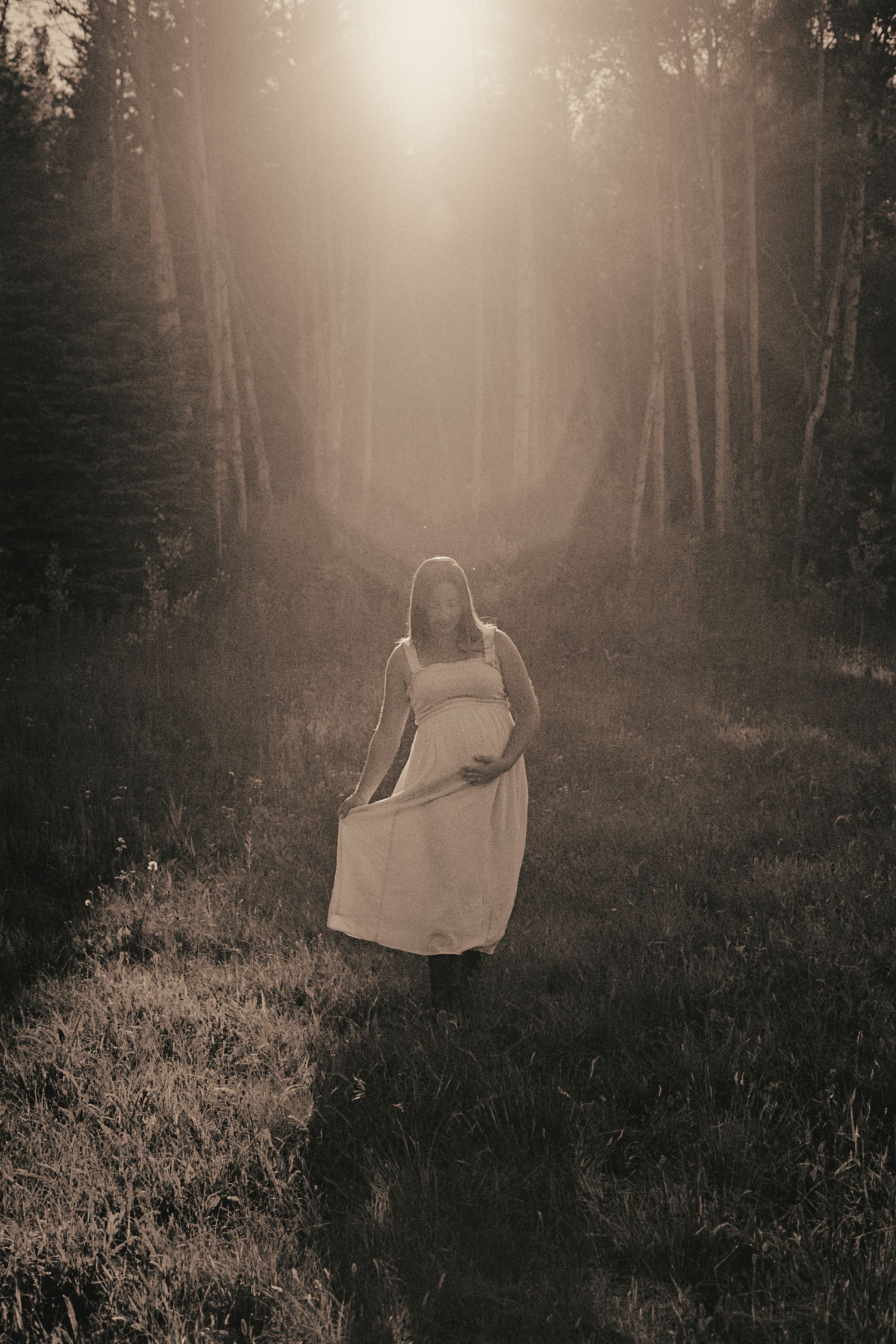 Ethereal sepia-toned photo of pregnant woman walking through sun-drenched forest path.