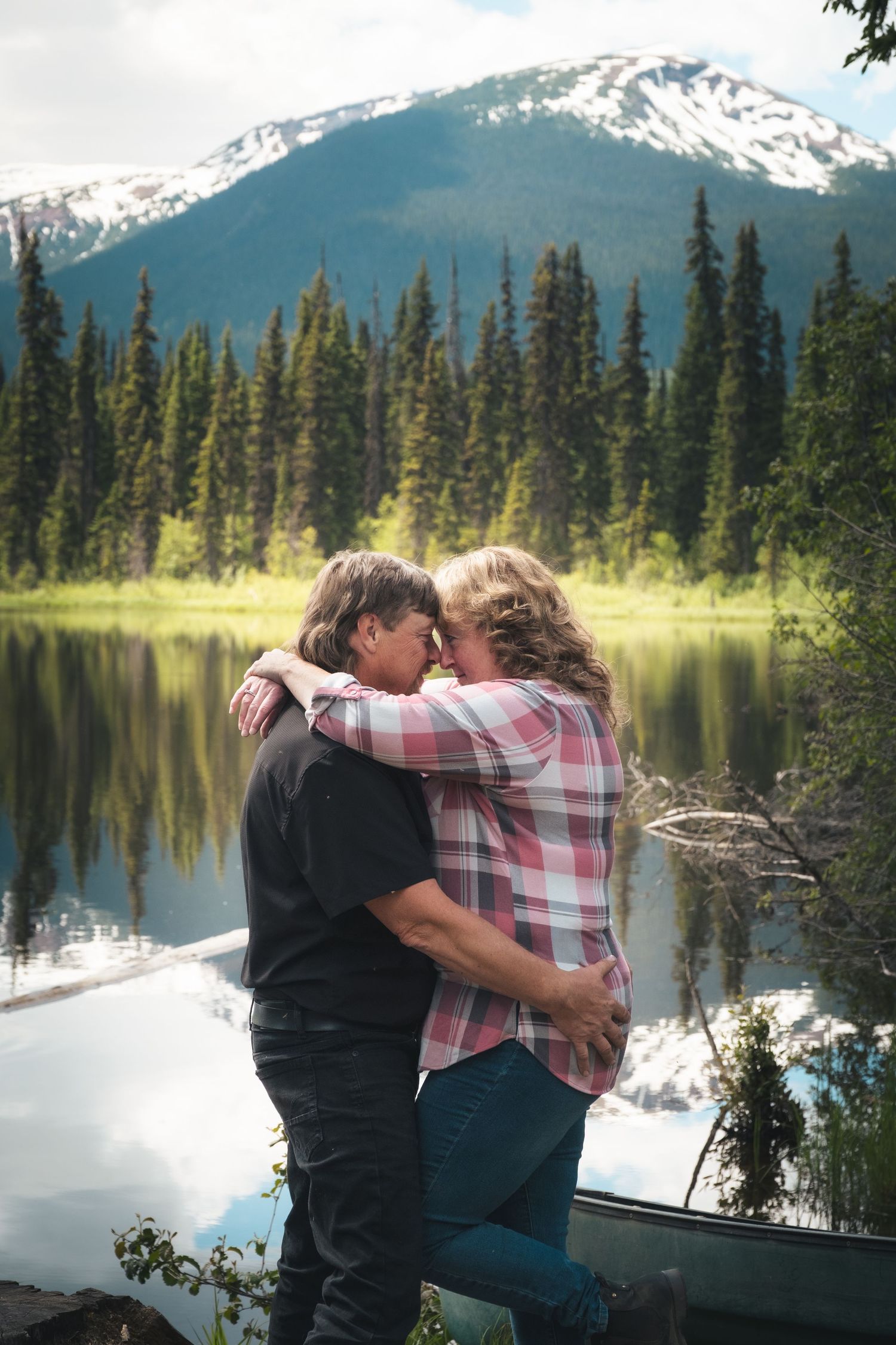 Couple sharing tender forehead touch embrace beside calm mountain lake with canoe nearby.