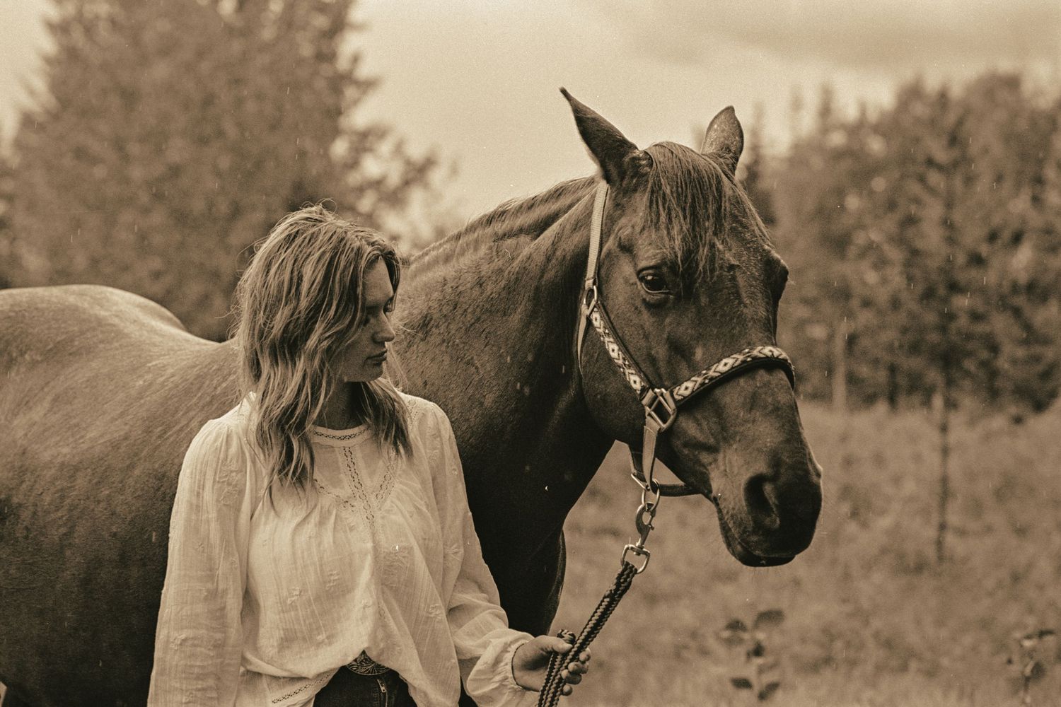 A young blonde woman in a white blouse stands beside a brown horse on a lead in a sepia-toned outdoor photo.
