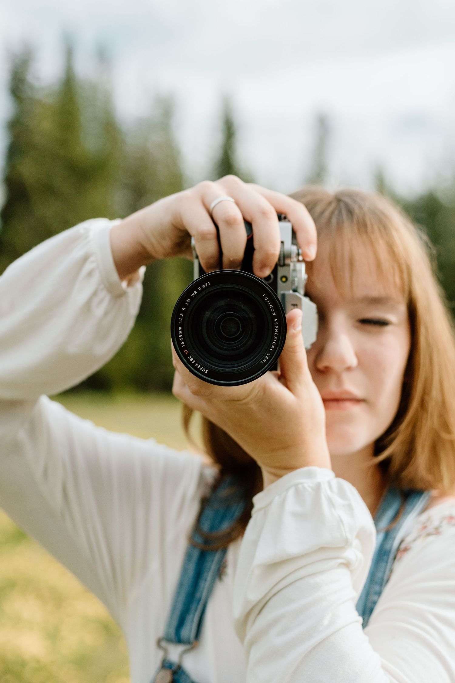 Young female photographer in denim overalls examines a camera lens outdoors in a green meadow.