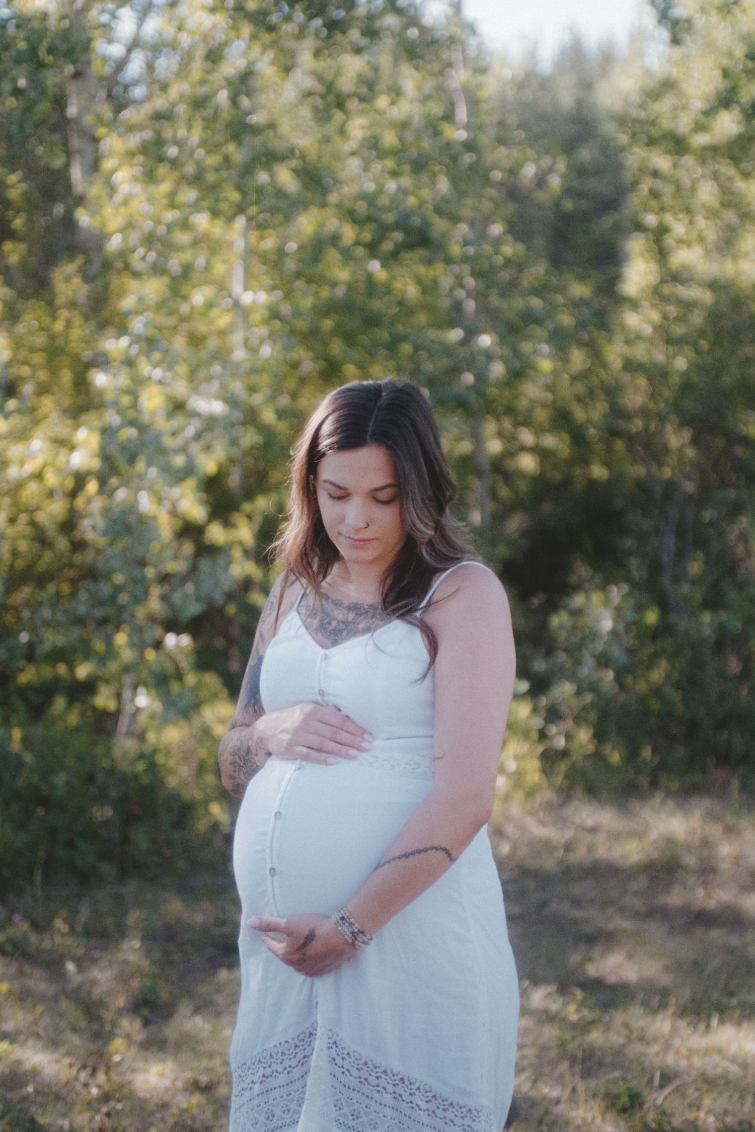 Pregnant woman in white lace dress standing outdoors among green trees, cradling her baby bump.