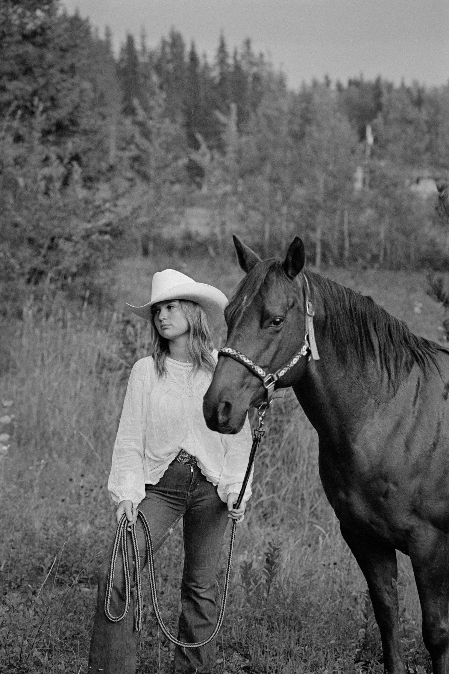 Black and white photo of a cowgirl in a white hat holding a rope beside a dark horse in a rural field.