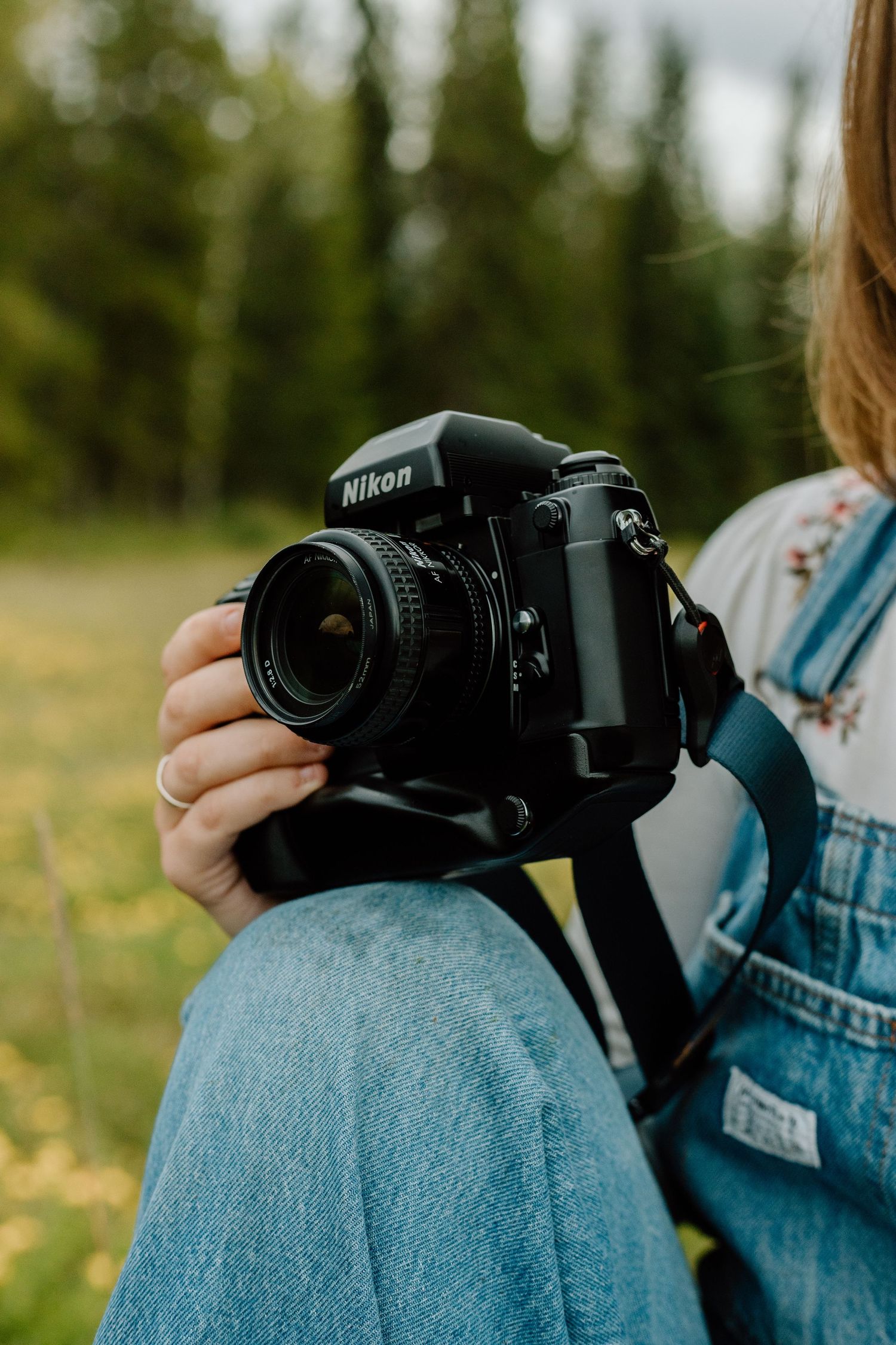 Close-up of a Nikon film camera held by a person wearing denim overalls in a forest setting.