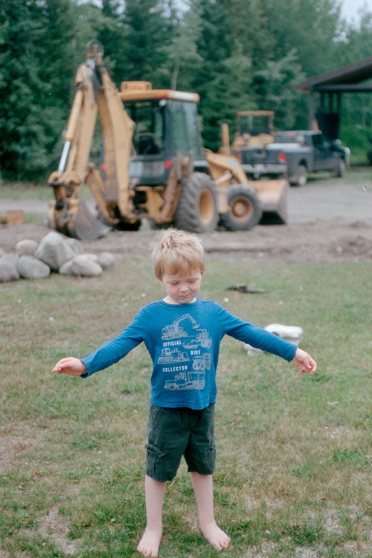 Young barefoot boy in blue shirt spreading arms wide with construction equipment in background.