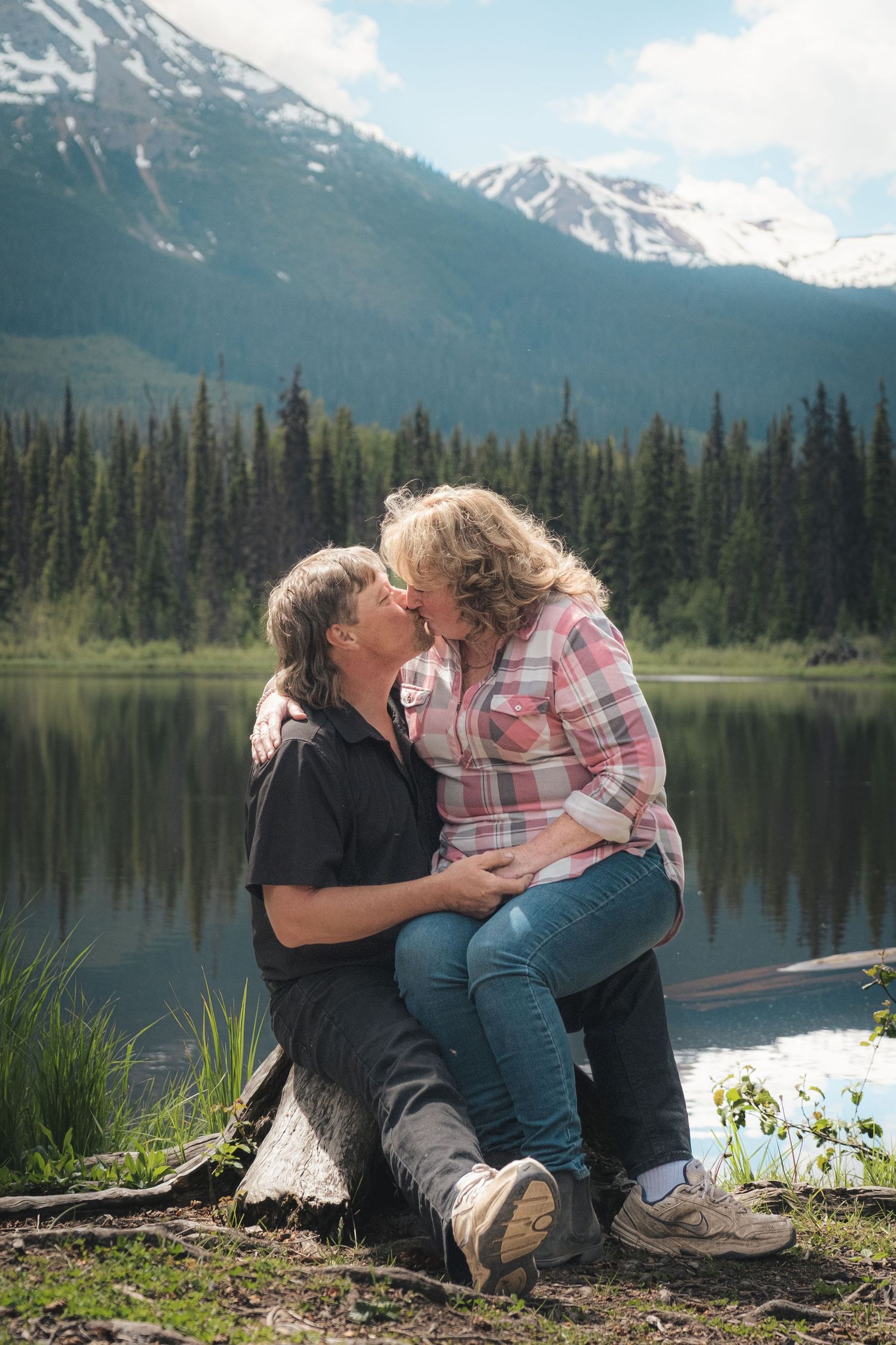 Couple sharing a romantic kiss while sitting on log beside a serene mountain lake.