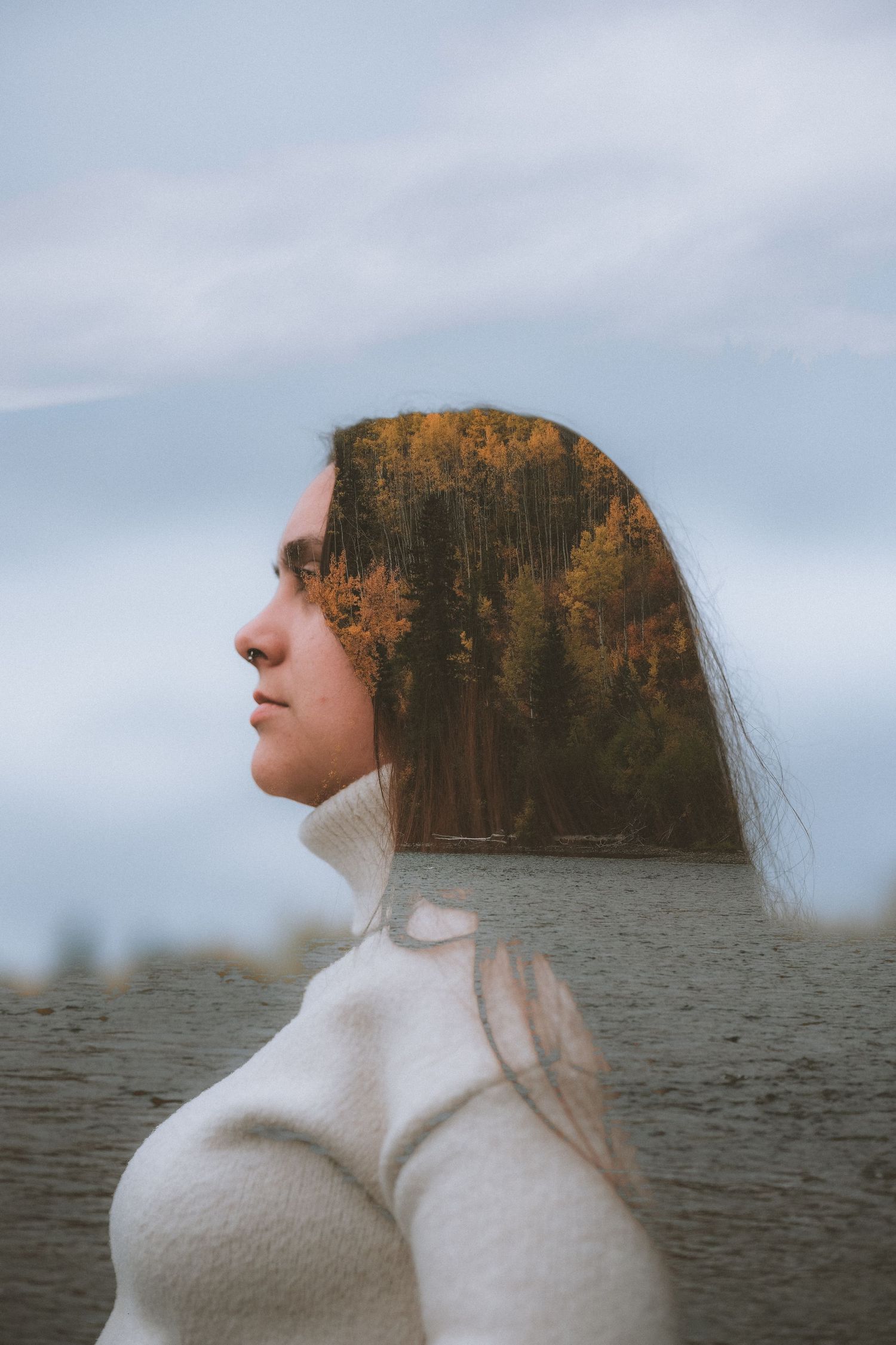 Double exposure portrait of a young woman in white turtleneck with autumn forest reflected on her profile.