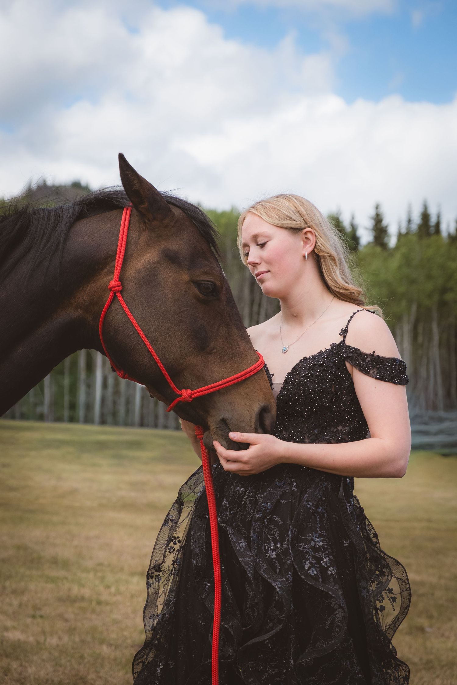 Young woman in black lace gown gently cradles dark horse's muzzle outdoors.