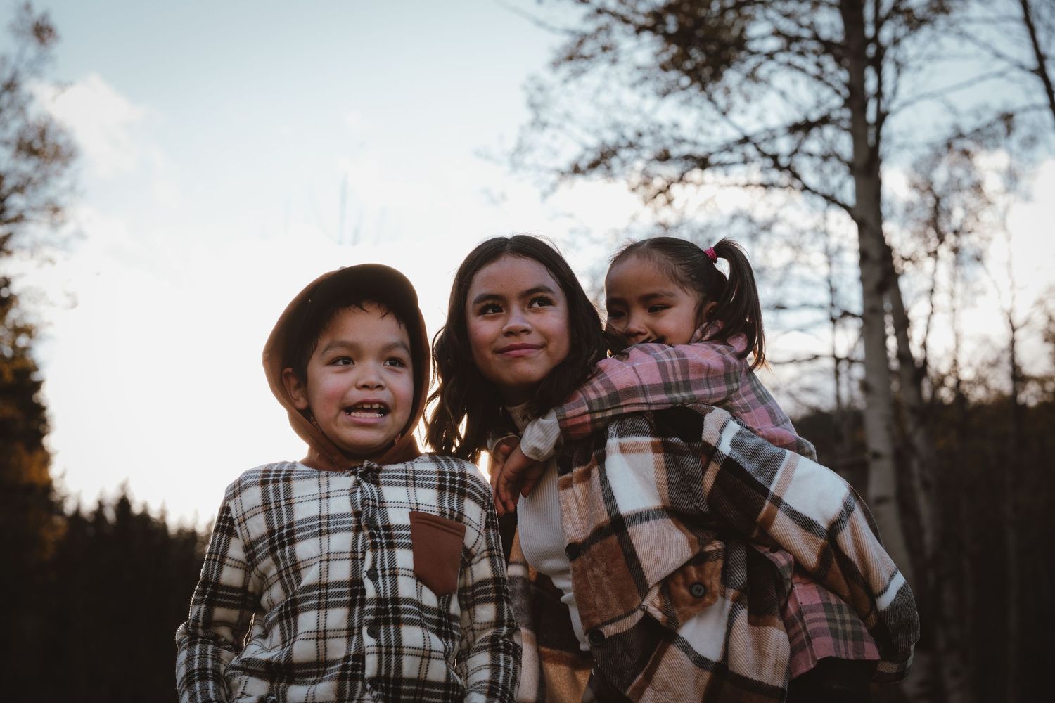 Three children in plaid outfits pose together in a forest at dusk during autumn.