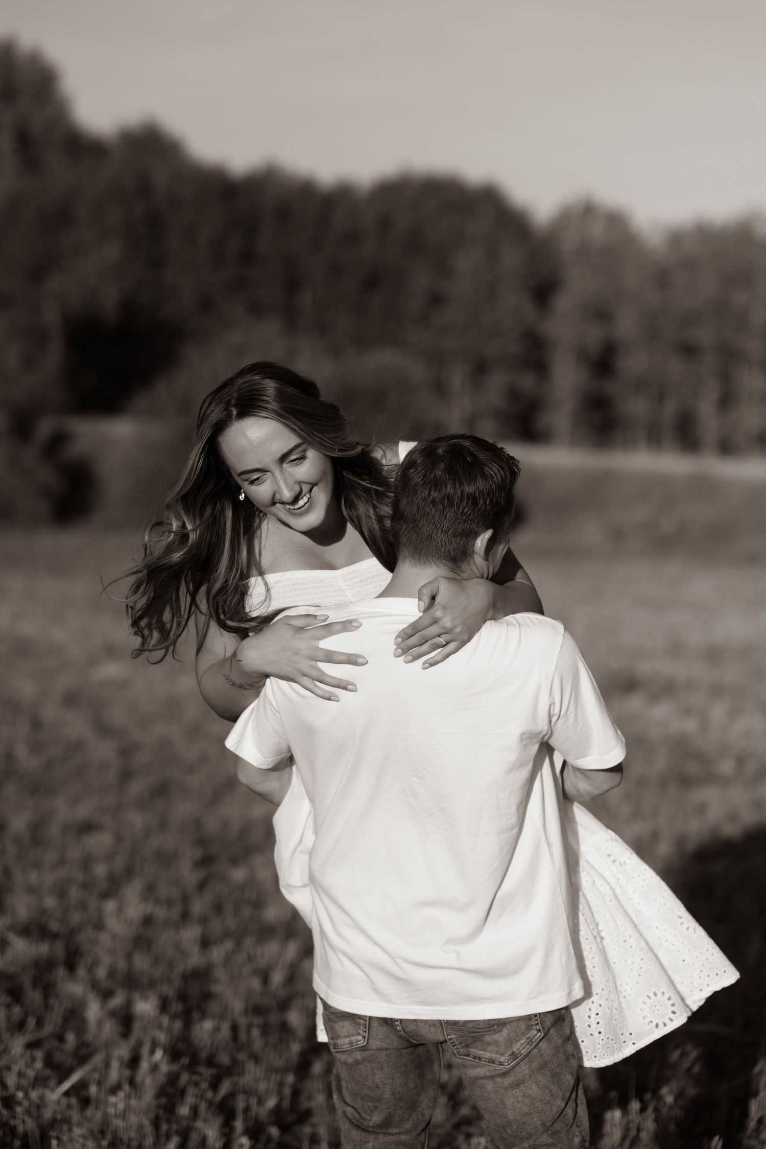 Black and white photo of a laughing couple embracing in an open field with trees in the background.