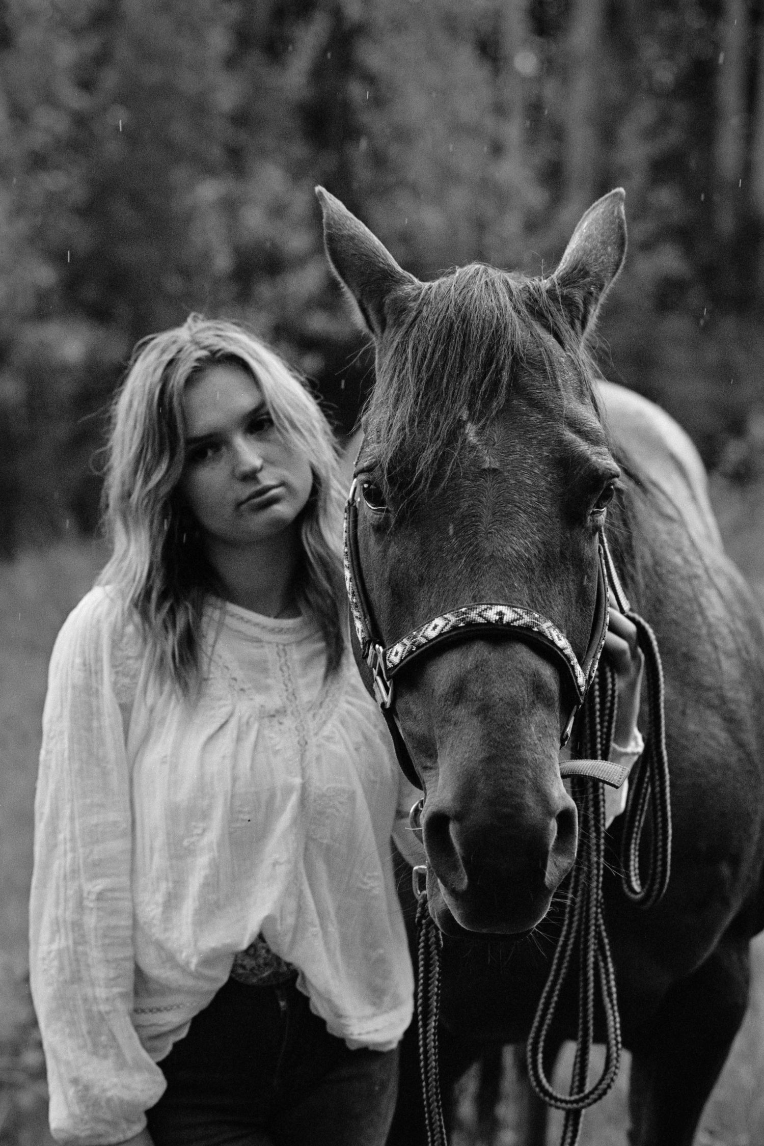 Black and white portrait of a young blonde woman standing beside a horse with a bridle in a forest setting.