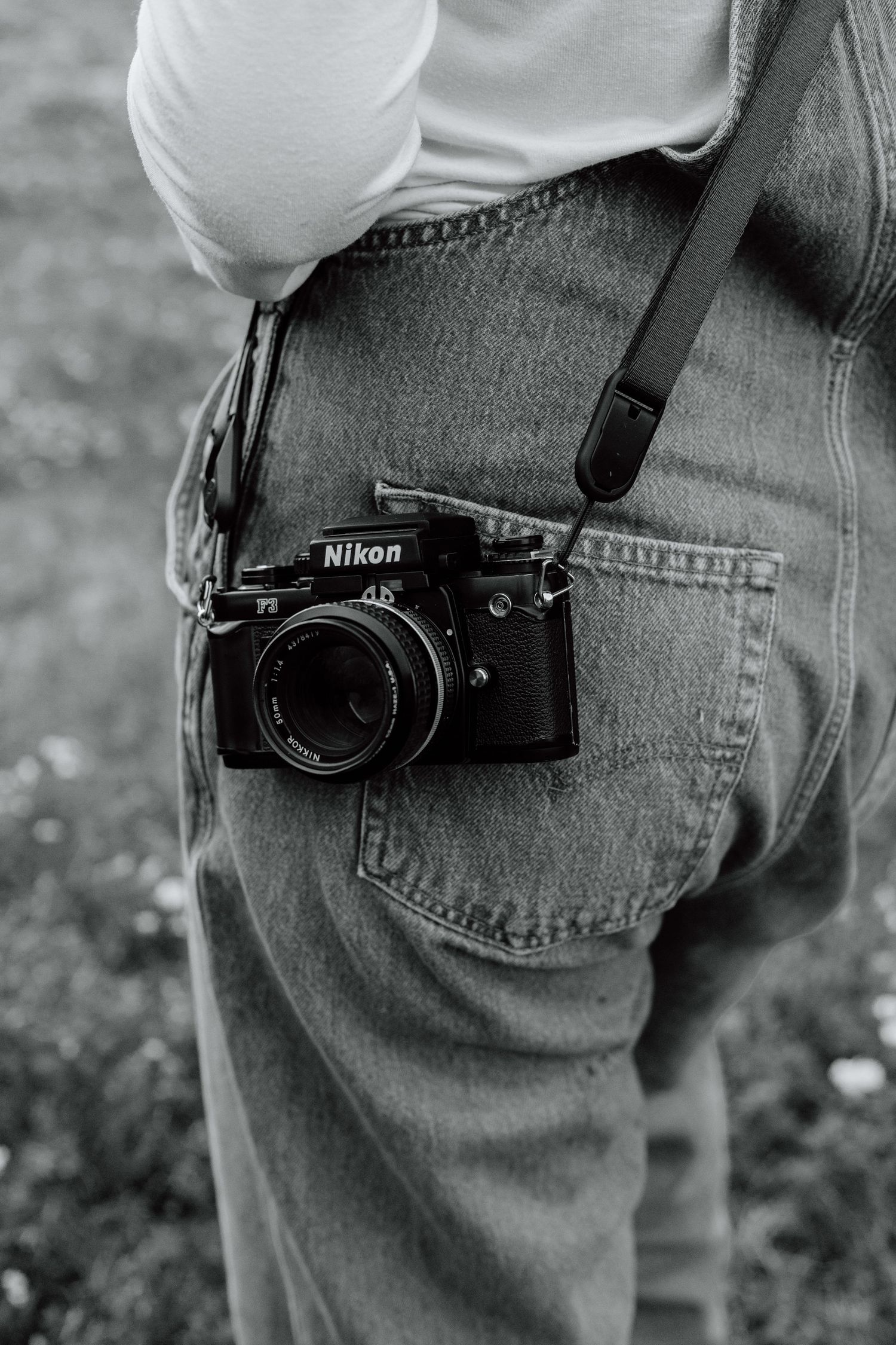 Black and white photo of a Nikon film camera tucked into denim overalls pocket with a strap.