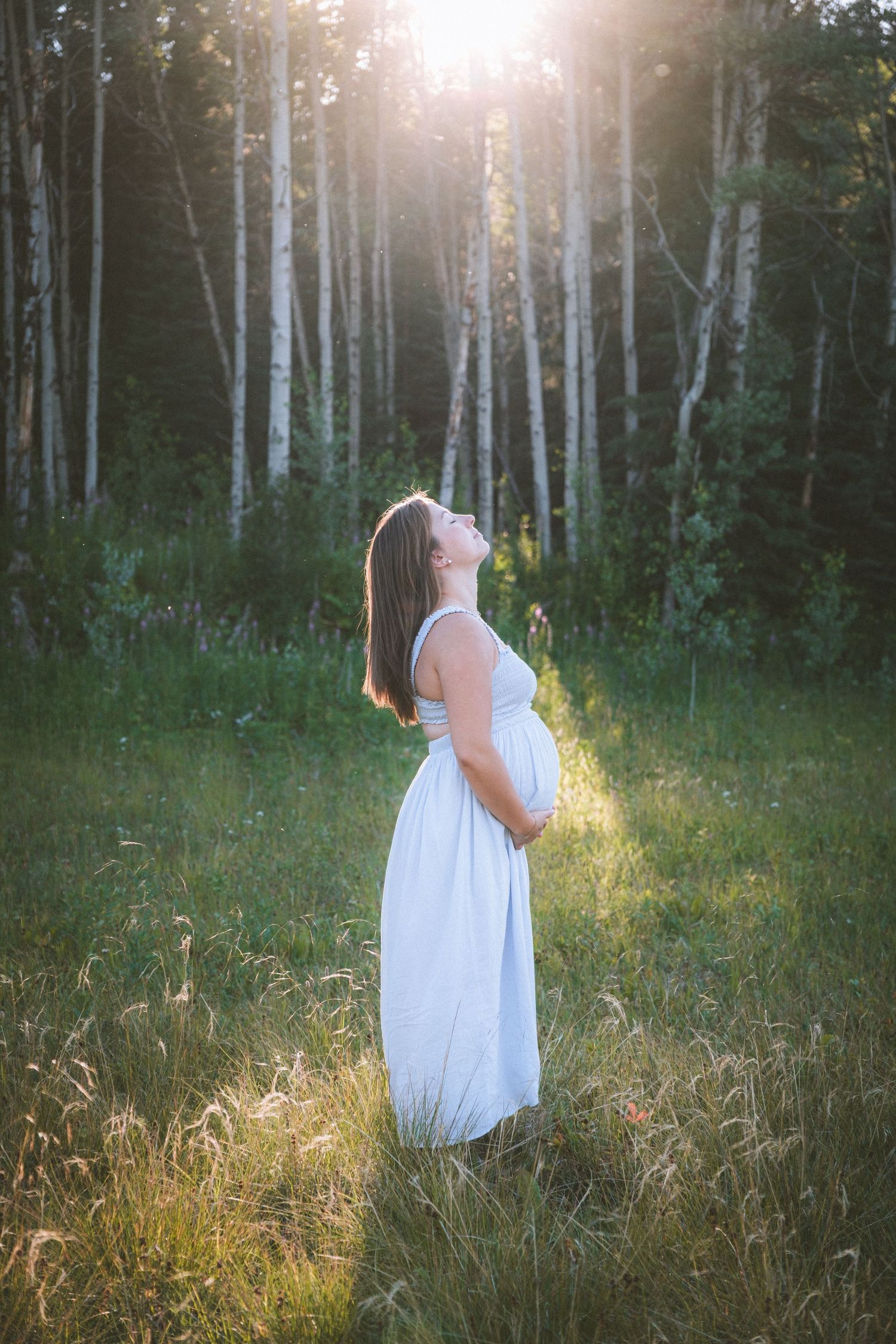 Expectant mother in white dress looking up at golden sunlight streaming through birch forest.