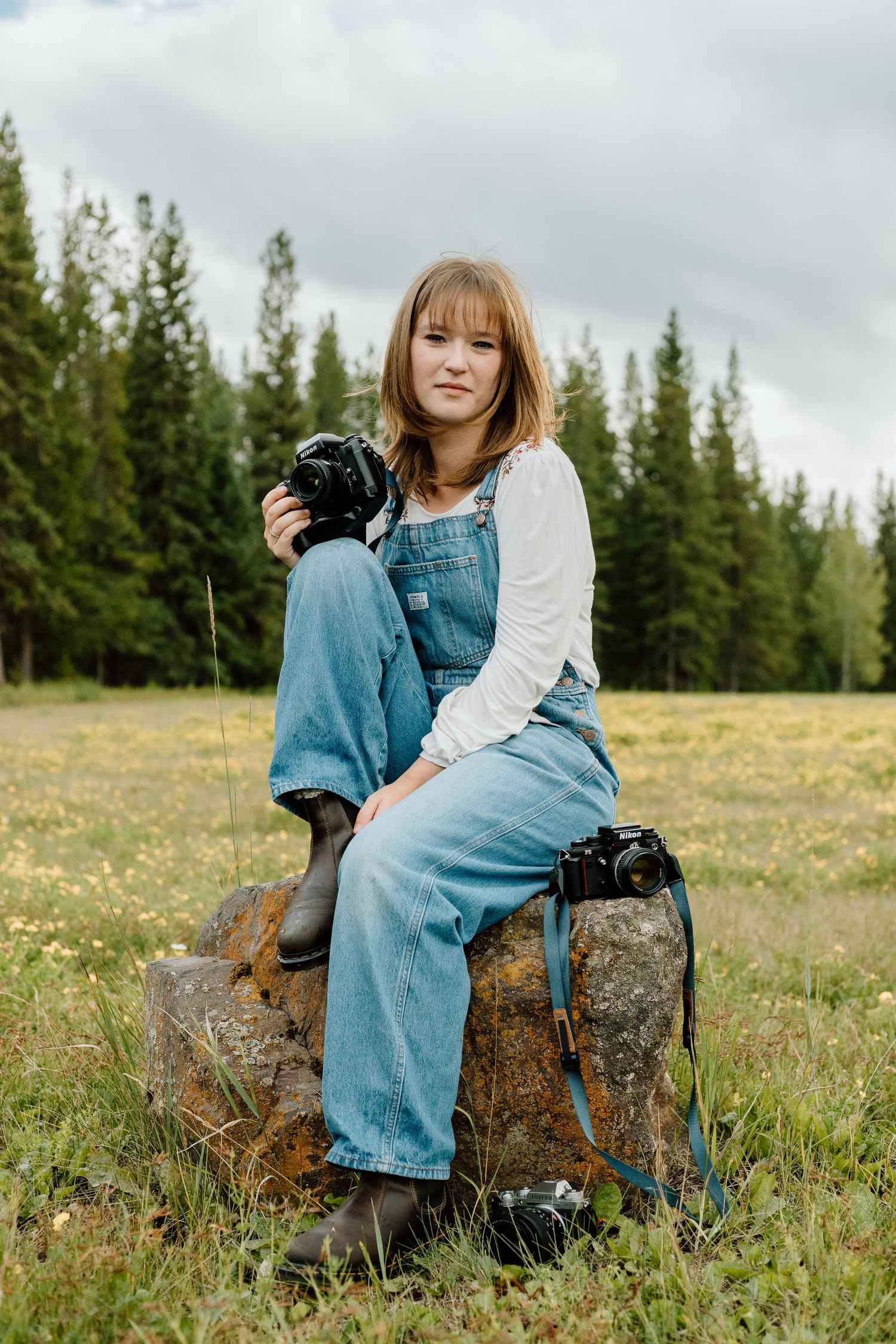 Young female photographer in denim overalls sits on a rock holding a Nikon camera in a meadow.
