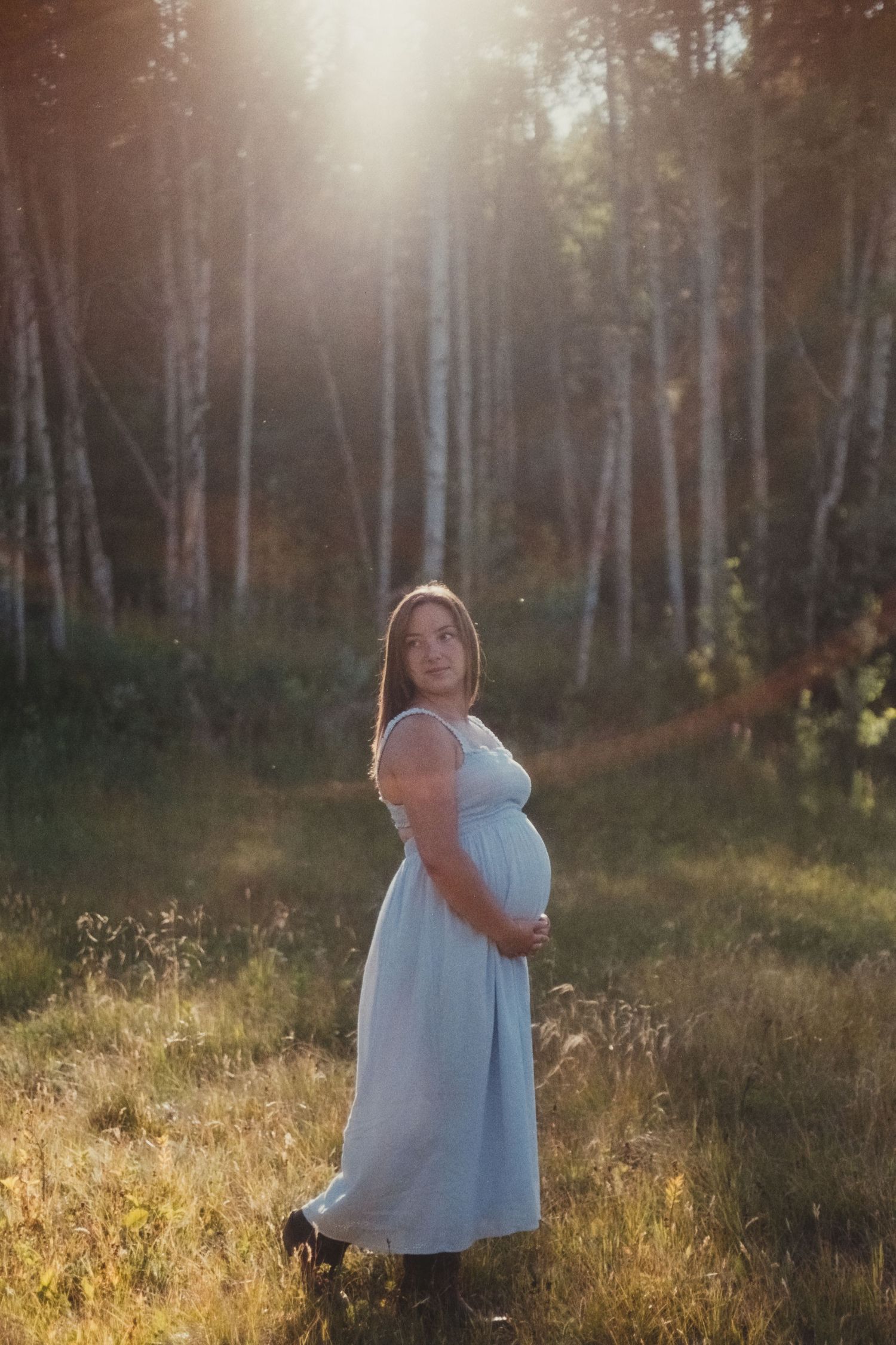 Pregnant woman in blue dress standing confidently in sunlit birch forest clearing.