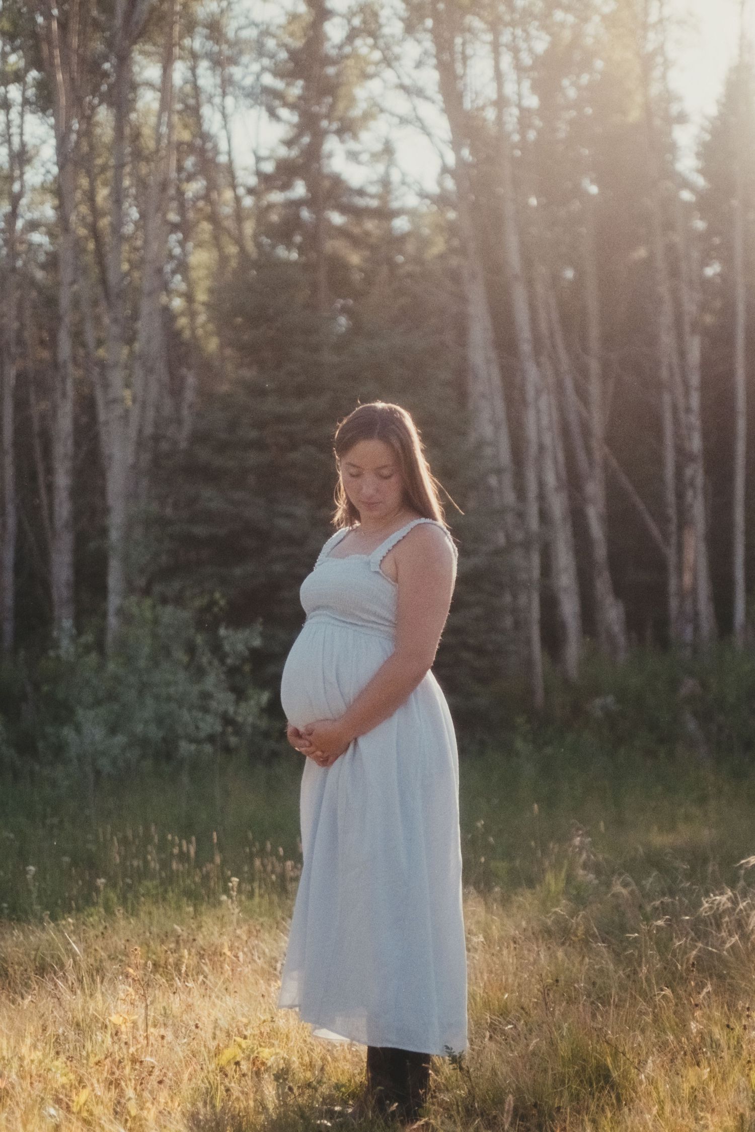 Pregnant woman in light blue dress cradling baby bump in sunlit forest clearing.