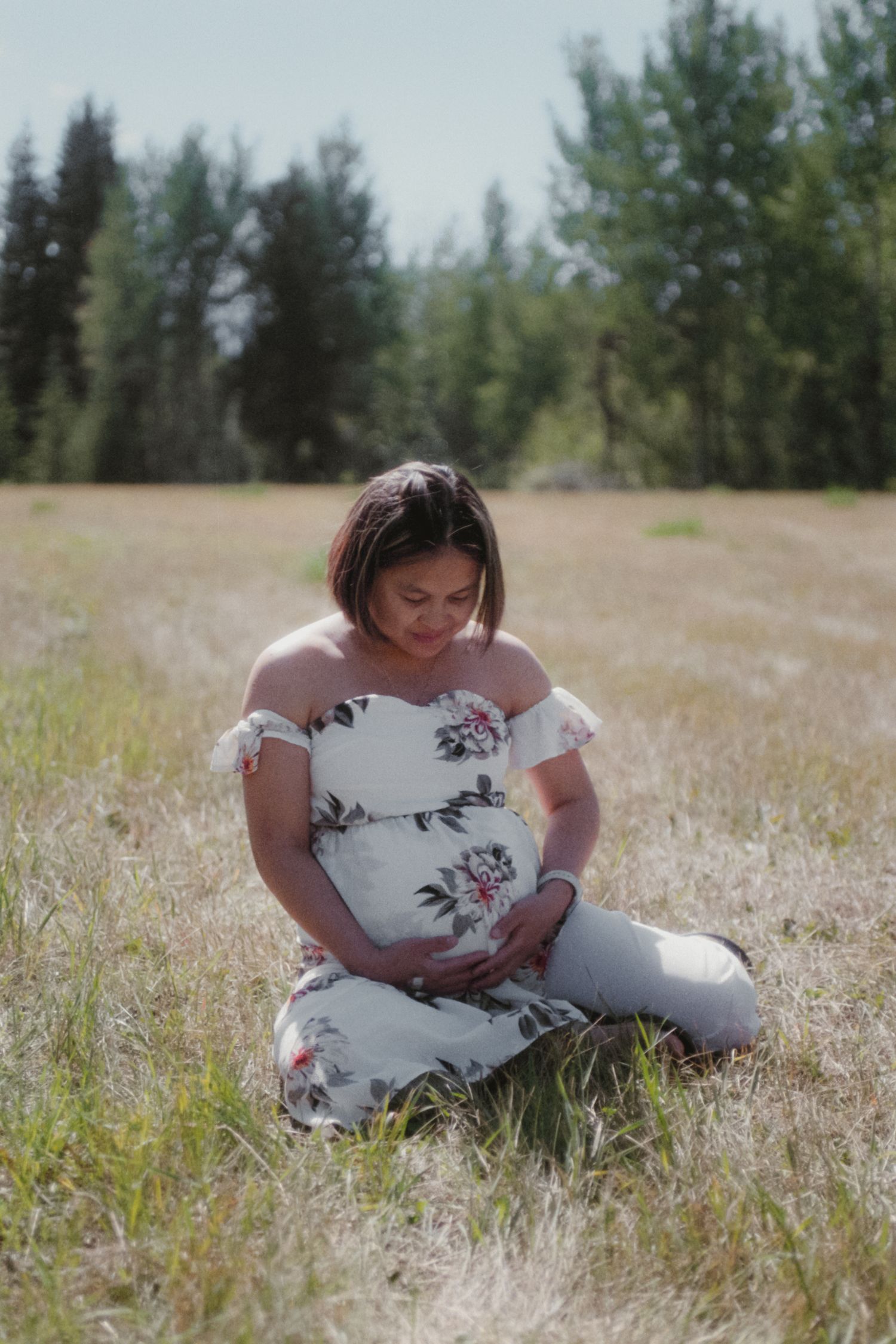 Pregnant woman in white floral dress sitting in a golden grass field, gently holding her baby bump.