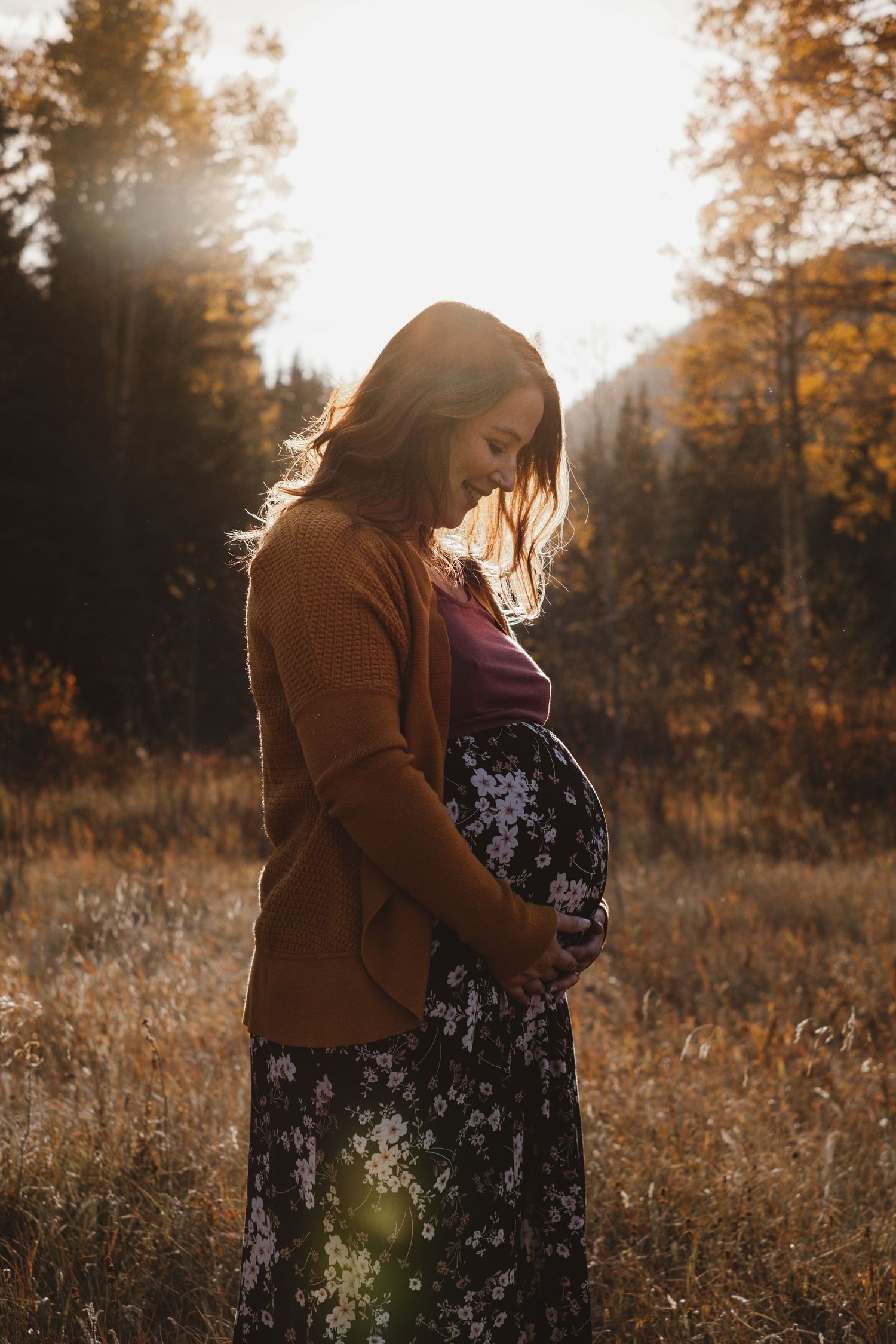Pregnant woman in rust cardigan cradling baby bump in a glowing autumn forest meadow.
