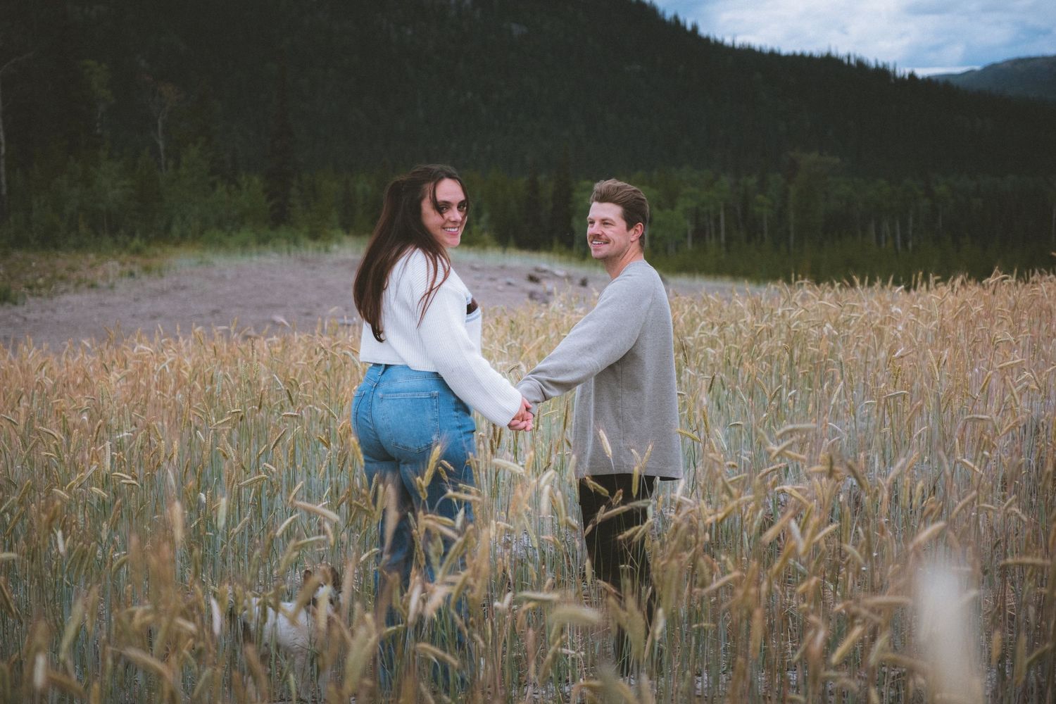 Smiling couple holds hands in a golden wheat field with forest and mountains in background.