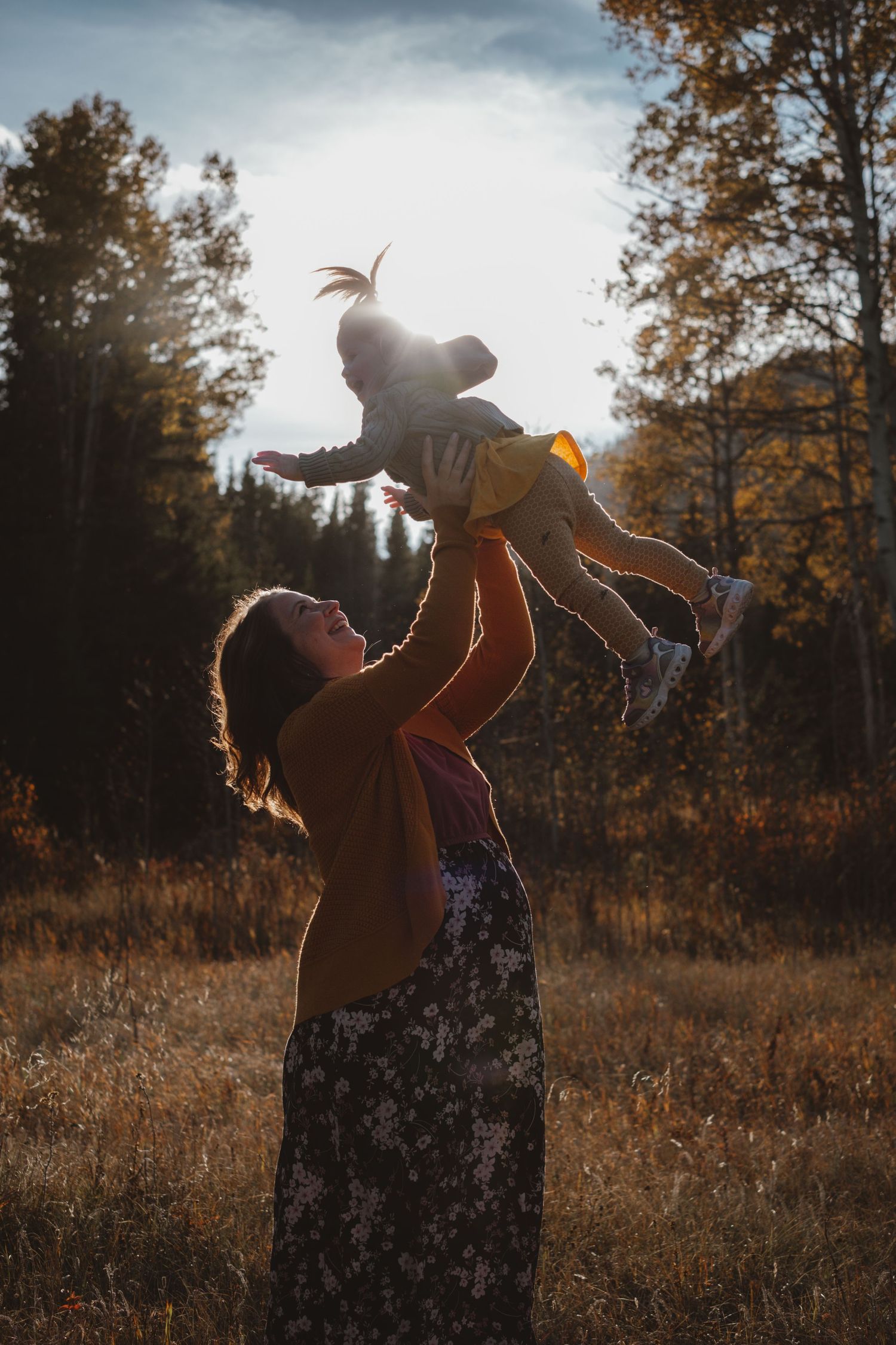 Mother joyfully lifting toddler into the air in a sunlit autumn meadow with golden trees.