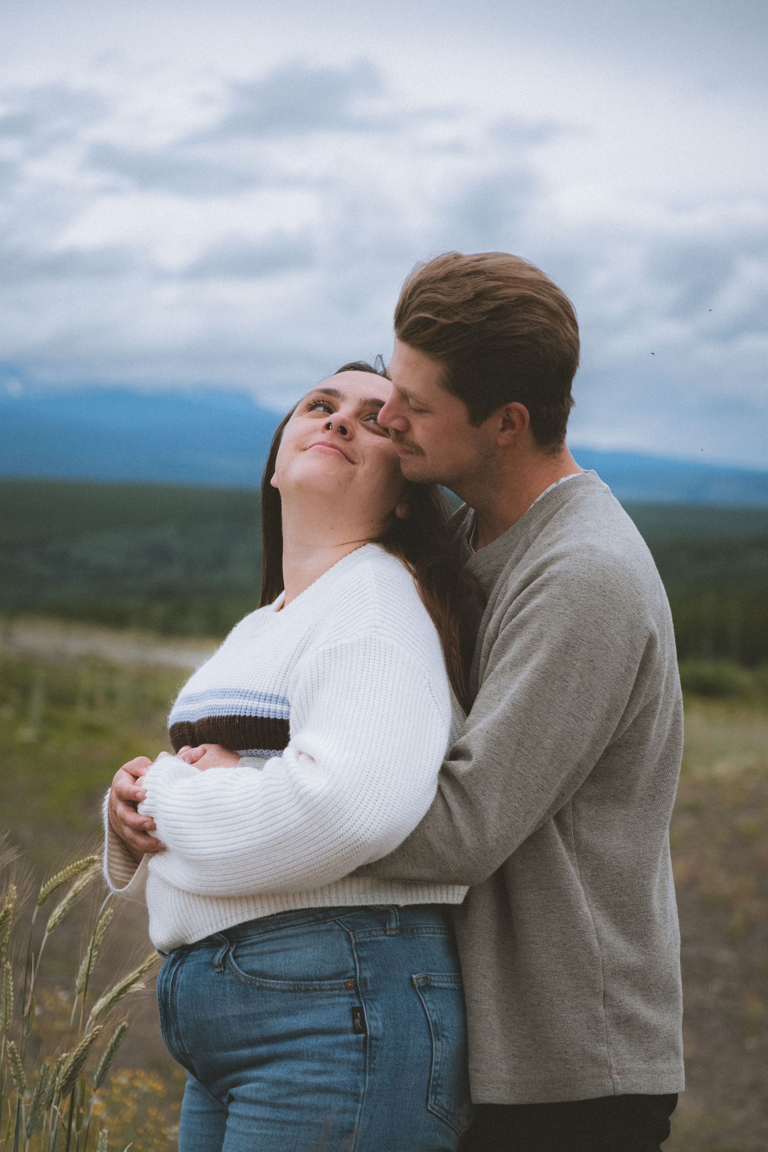 Man kisses woman on cheek as she smiles with eyes closed in a scenic mountain field.