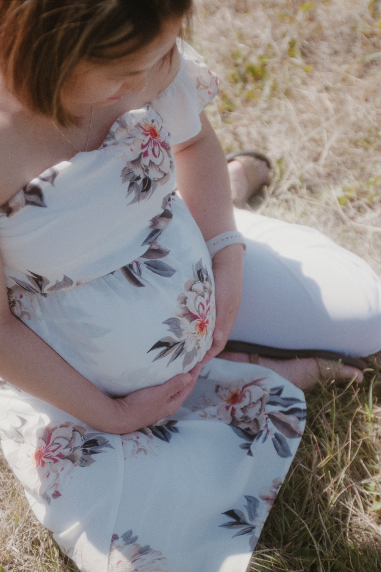 Close-up of pregnant woman in white floral off-shoulder dress cradling her baby bump outdoors.