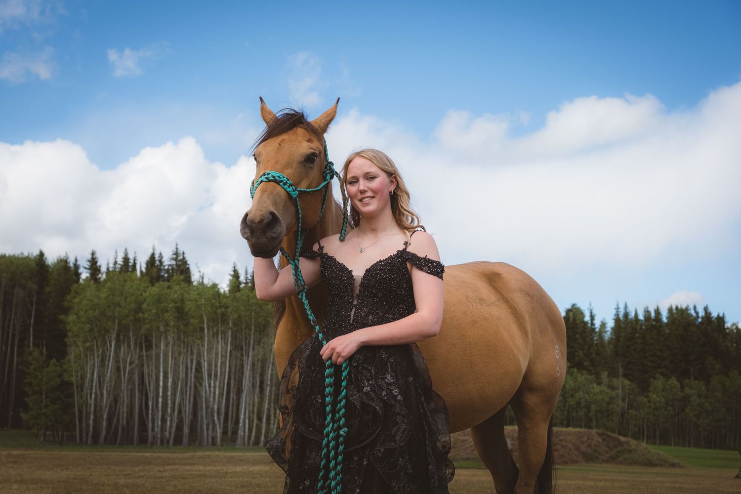 Smiling blonde in black lace dress poses beside brown horse in sunny field.