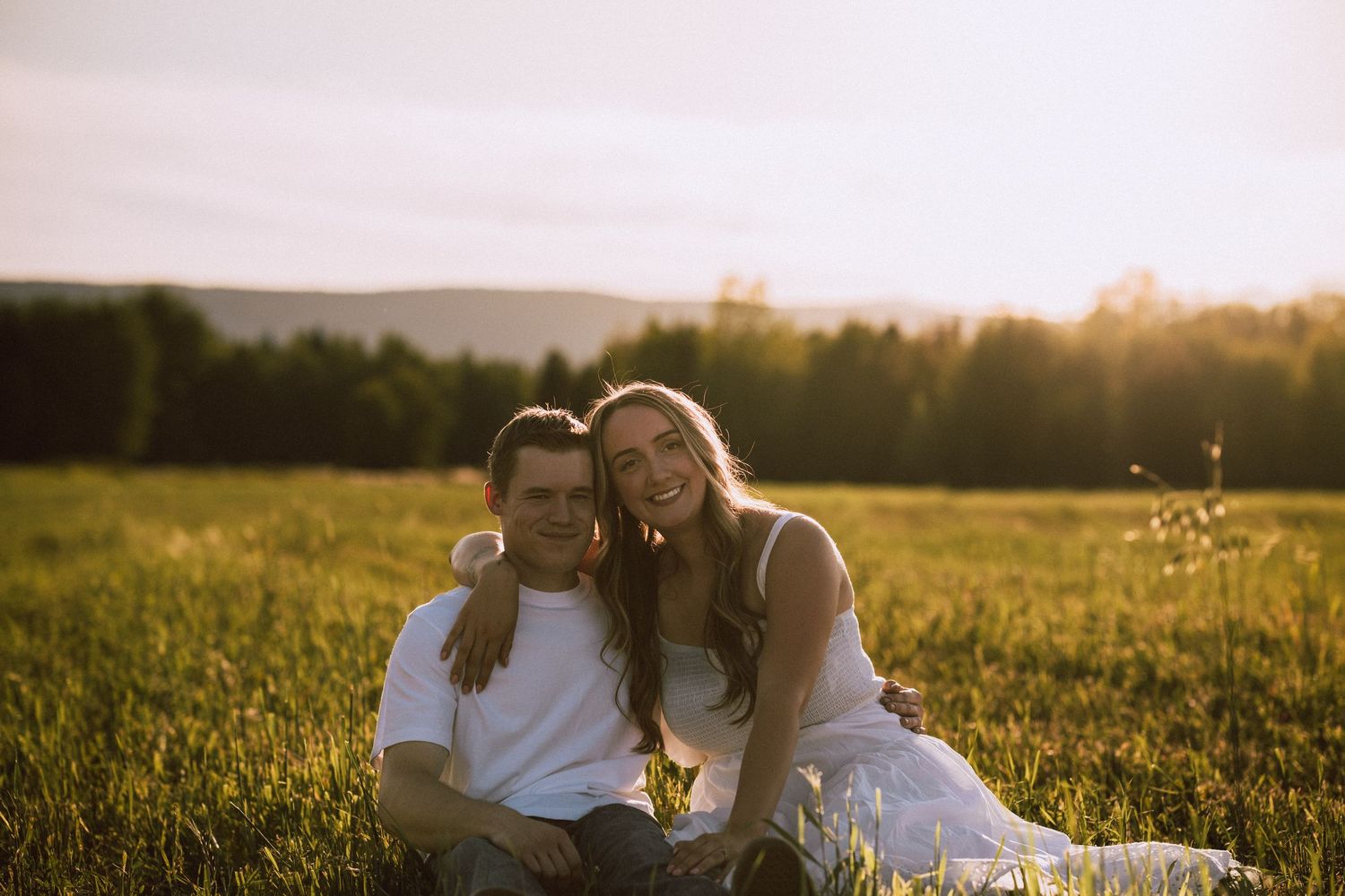 Happy couple sitting together in a sunlit green meadow smiling warmly during a golden hour photo session.