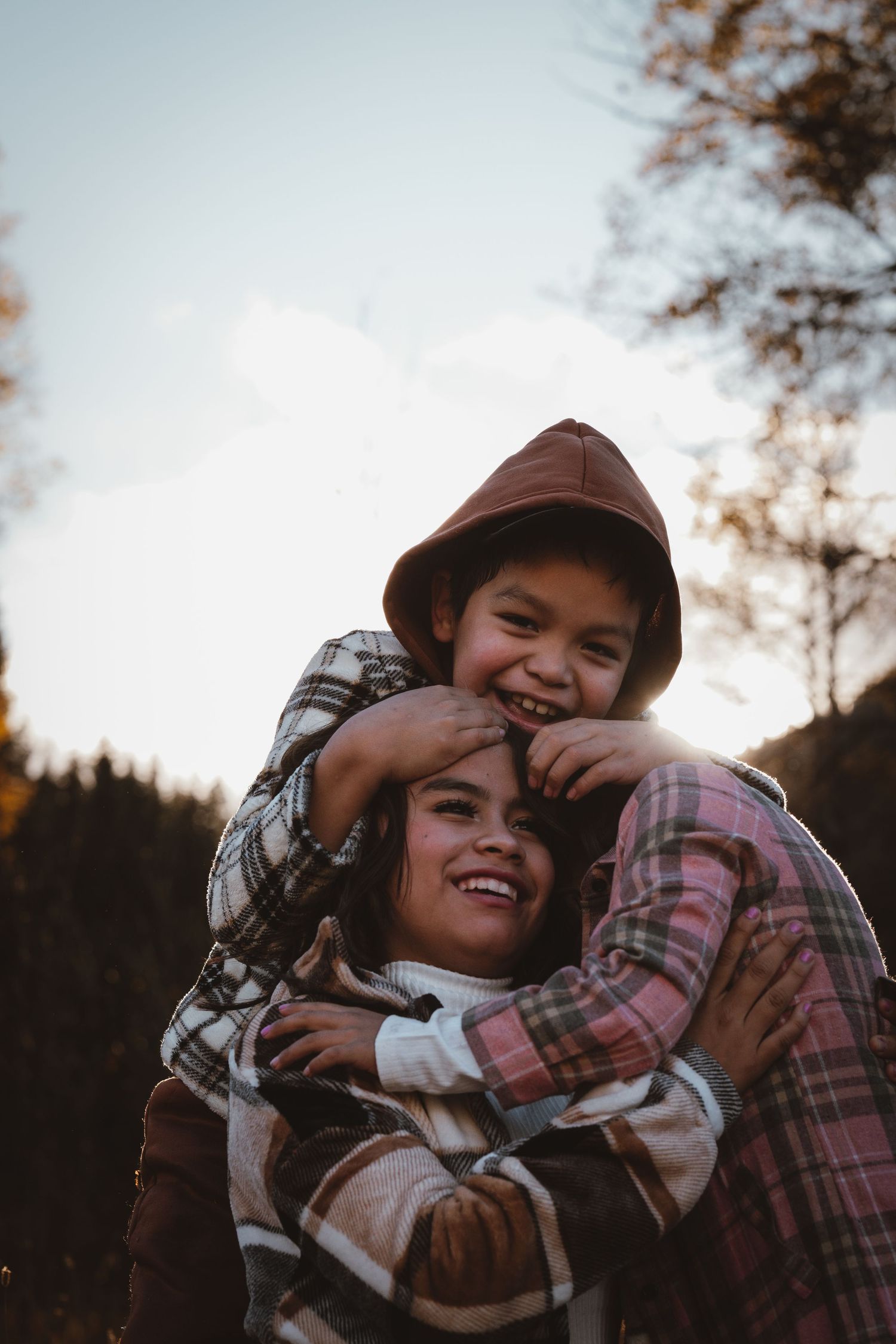 A boy in a brown hood joyfully hugs a smiling woman in a plaid coat outdoors at sunset.