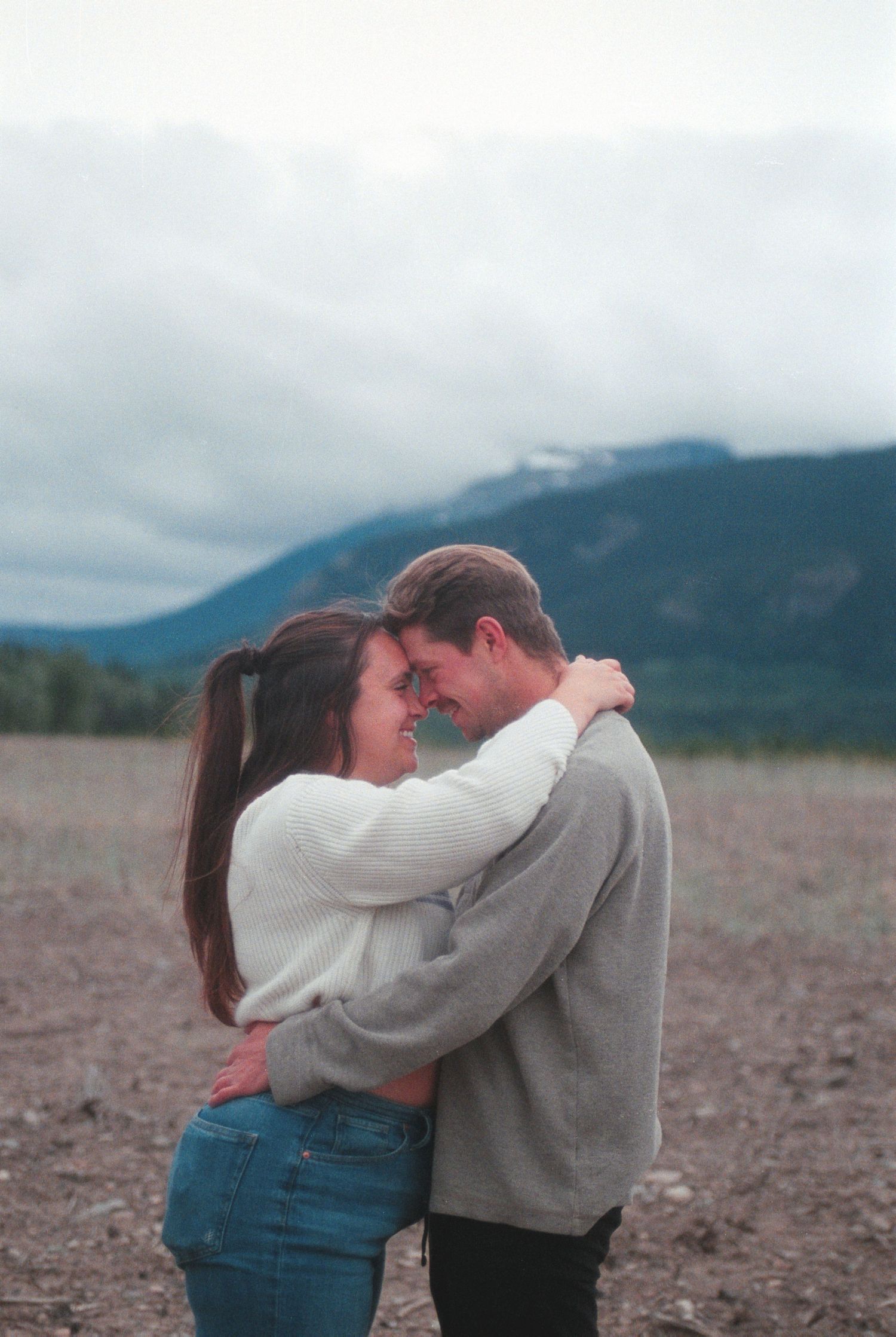 Loving couple embracing and smiling forehead to forehead in an open field with mountain backdrop.