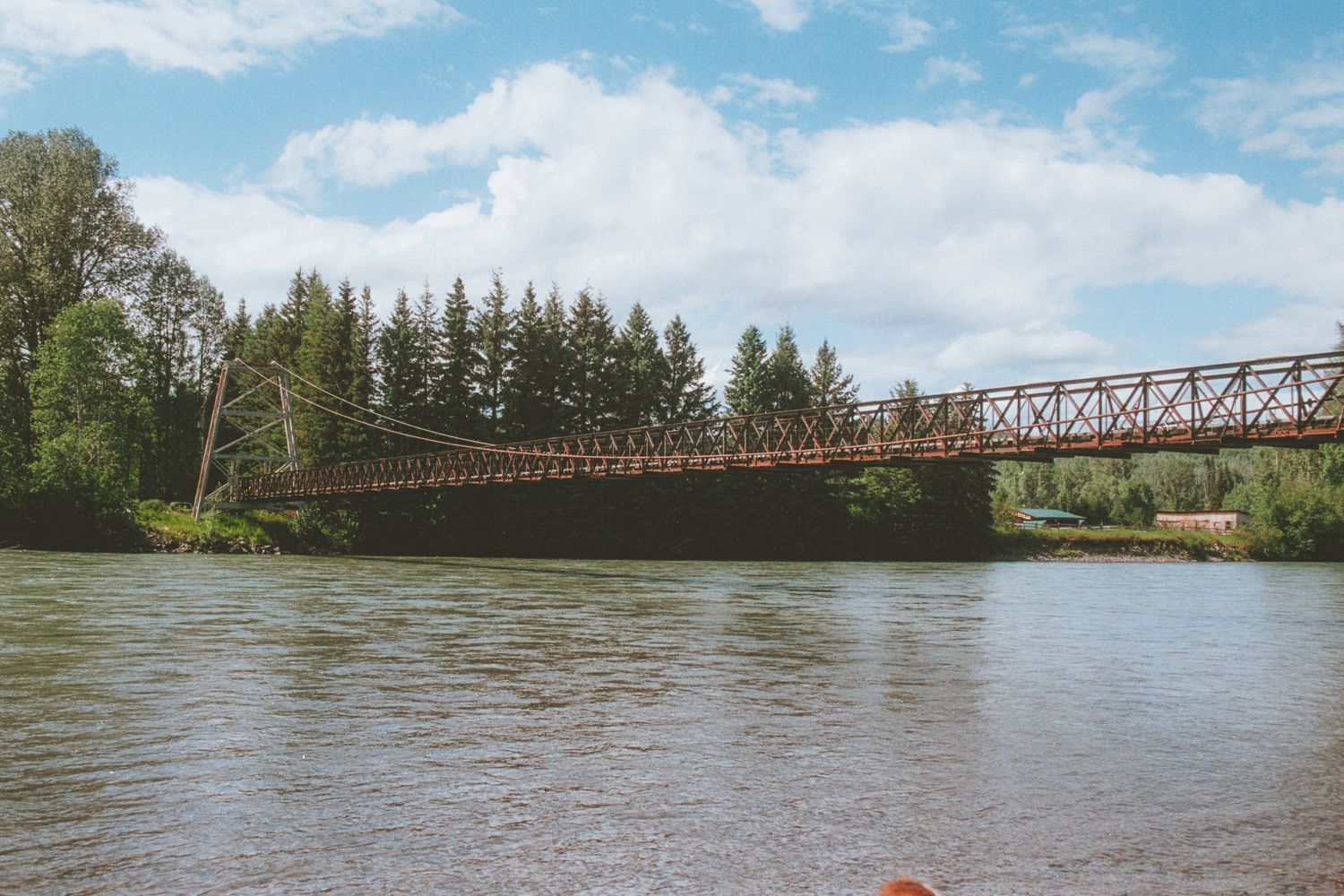 A rustic iron bridge spans a calm river surrounded by lush green trees under a partly cloudy sky.